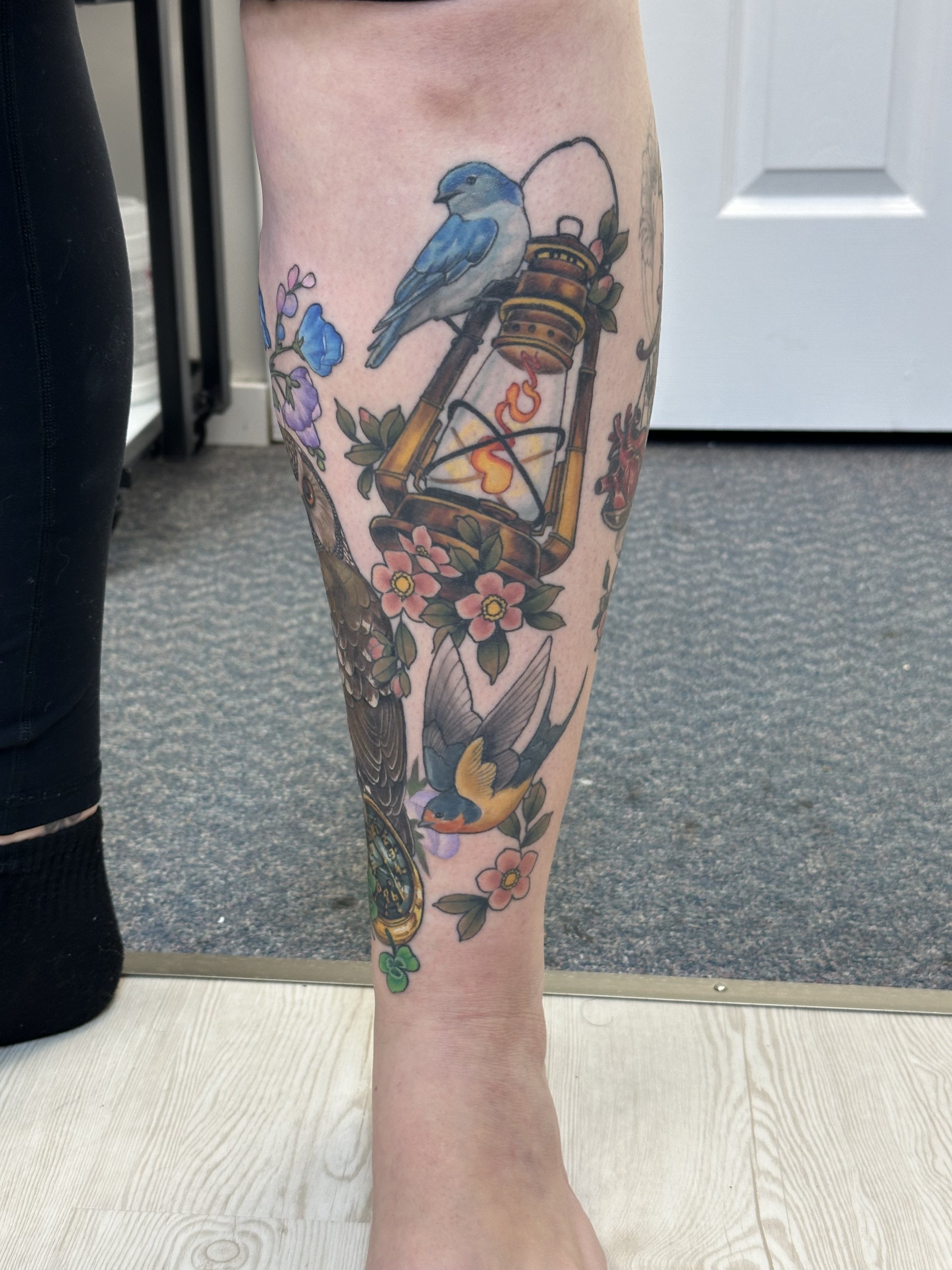 Leg tattoo featuring a blue bird on a lantern, surrounded by flowers and a swallow.