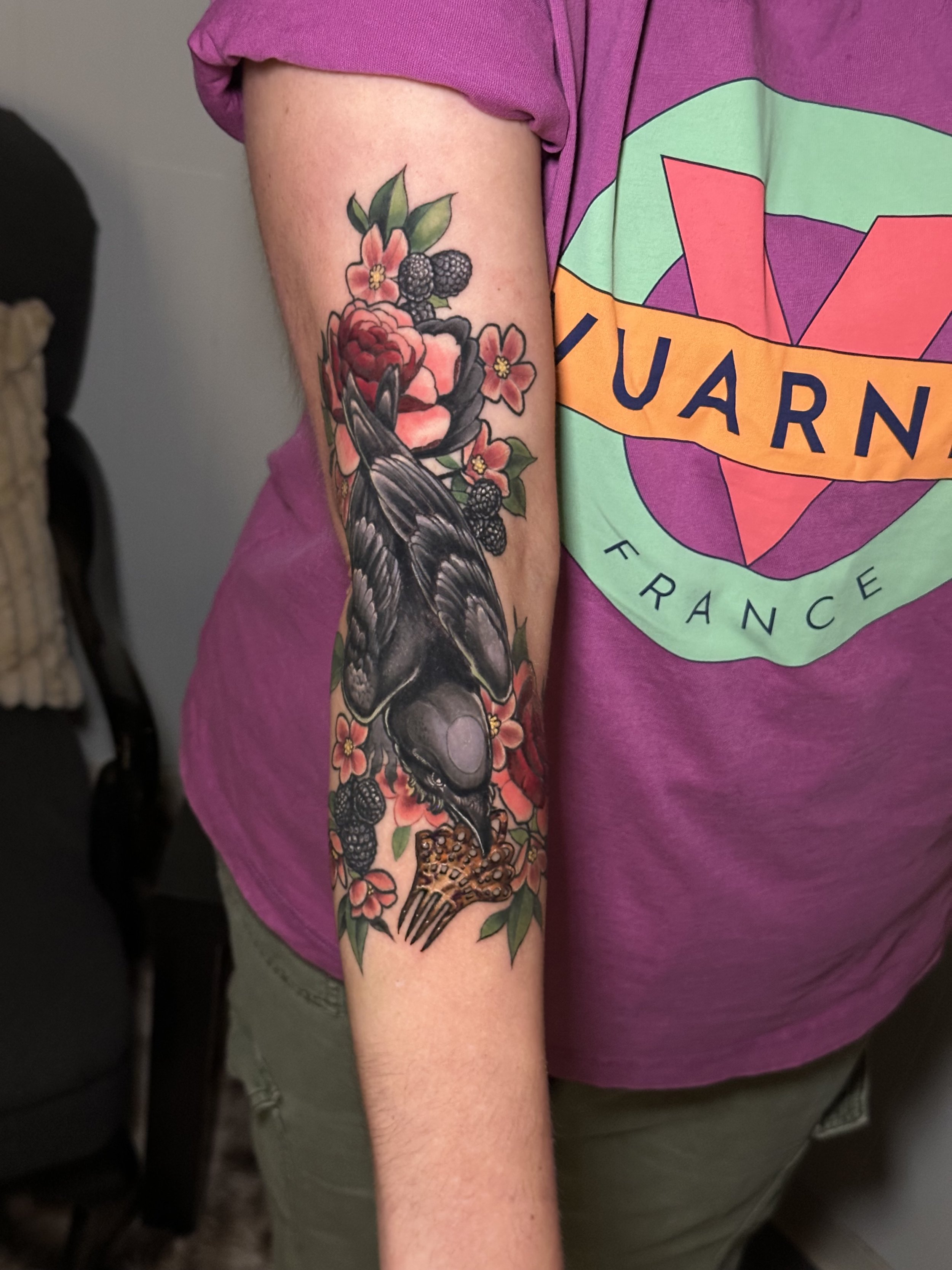 Person's forearm with a colorful tattoo of a raven, flowers, and blackberries, wearing a purple shirt with a logo.