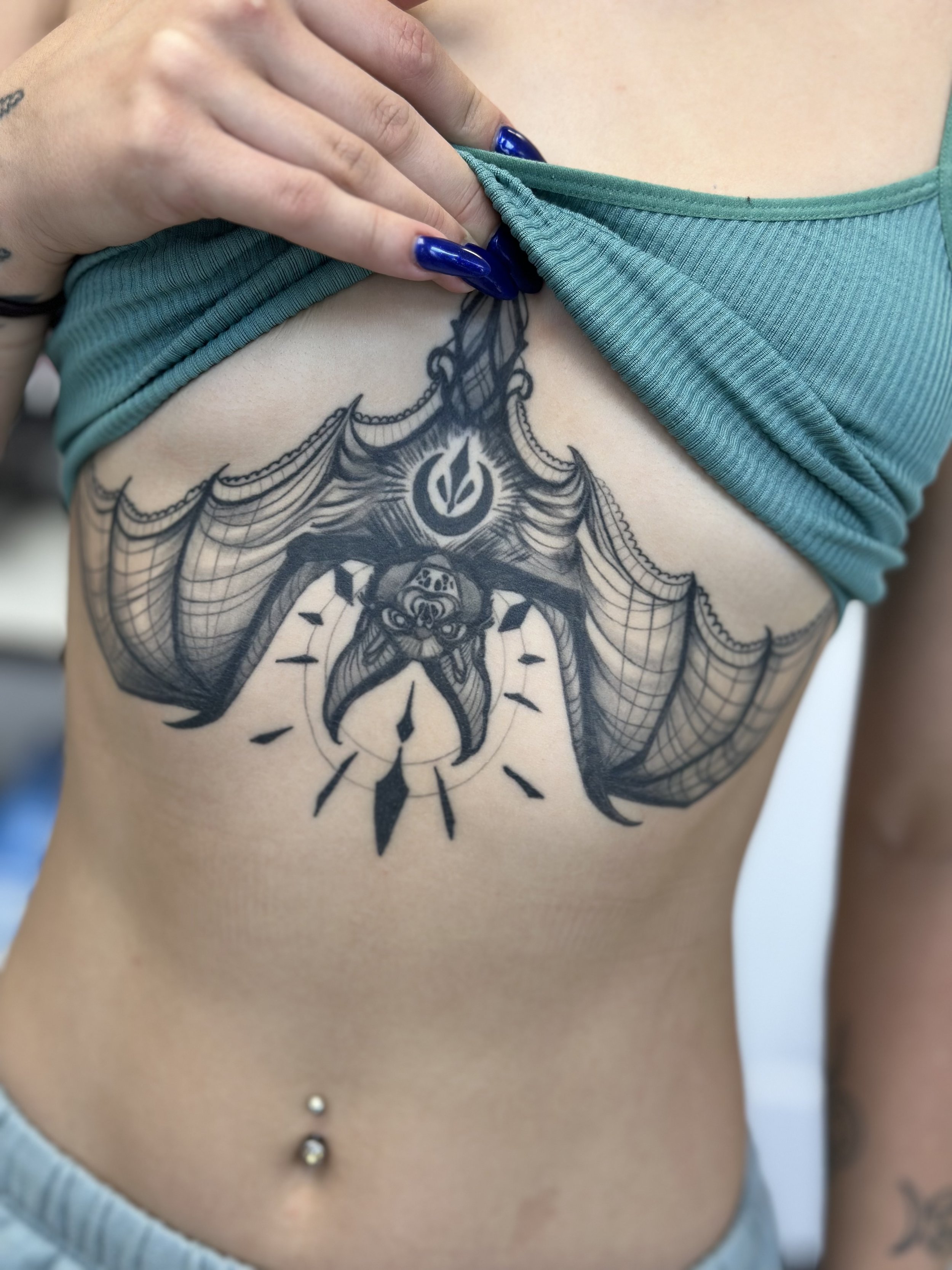 Large black and grey underboob tattoo featuring a symmetrical design with intricate details and a central artistic motif. The person is wearing a green top with blue nail polish visible.