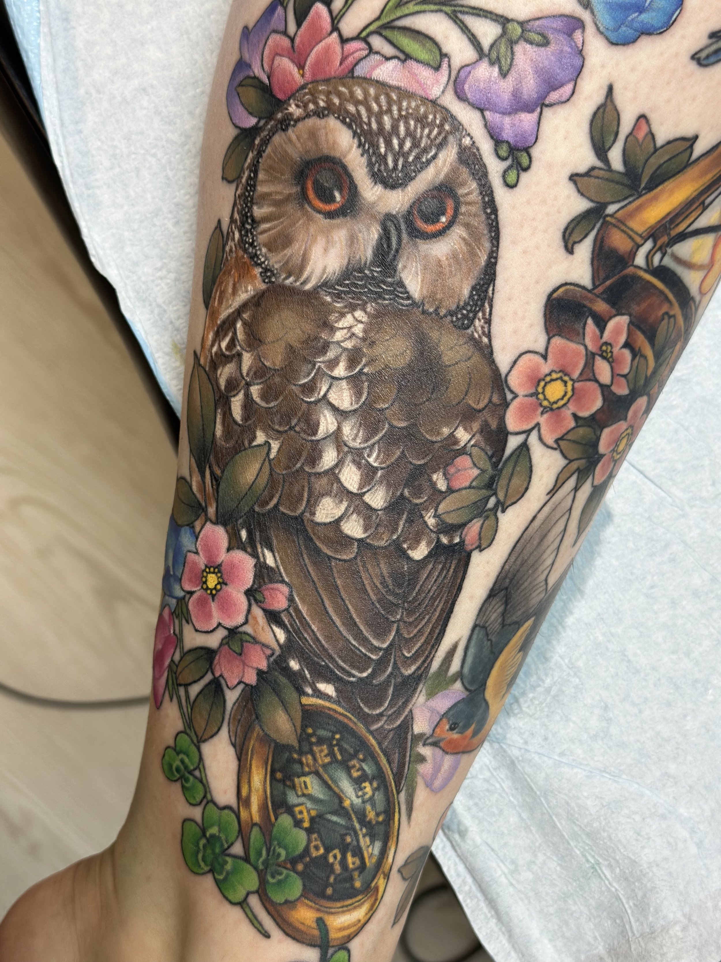 A tattoo featuring a detailed owl surrounded by colorful flowers and green leaves, with a pocket watch incorporated into the design.