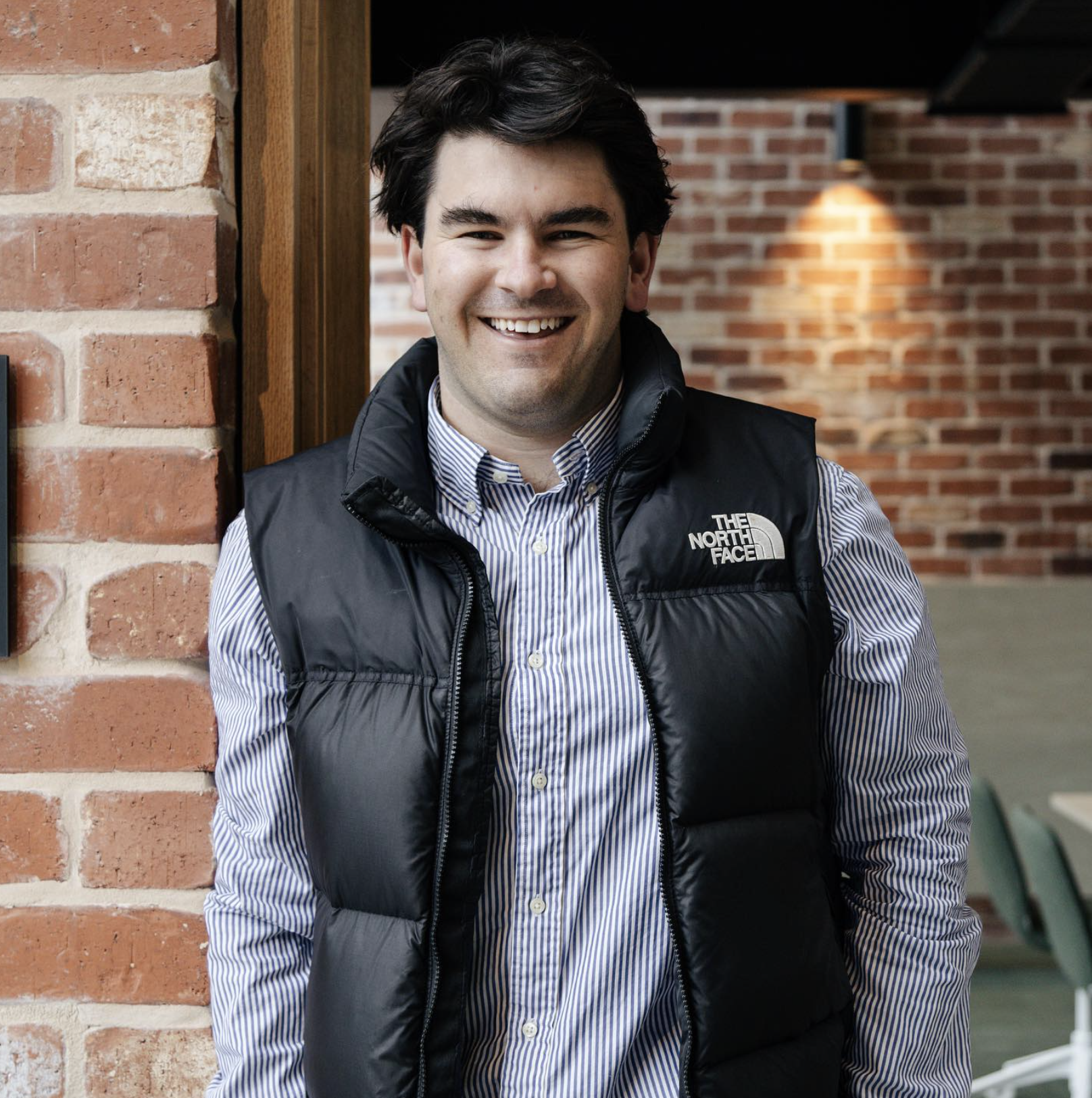 A smiling man with dark hair, wearing a striped button-up shirt and a black The North Face puffer vest, standing indoors against a brick wall with warm lighting.
