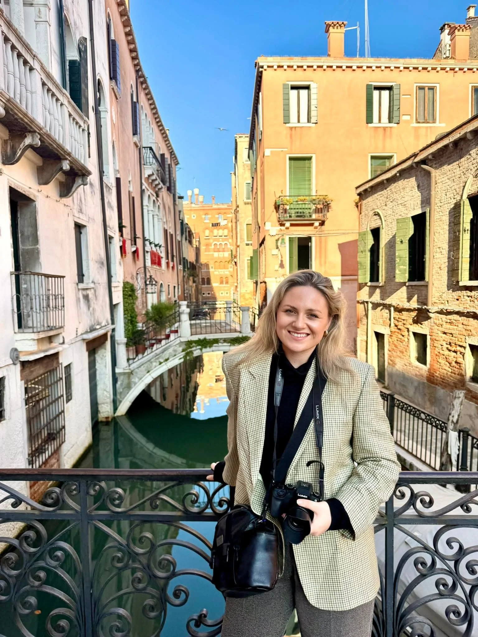 A smiling woman standing on a bridge over a canal in Venice, Italy, holding a camera with a camera strap around her neck, with colorful historic buildings and a blue sky in the background.
