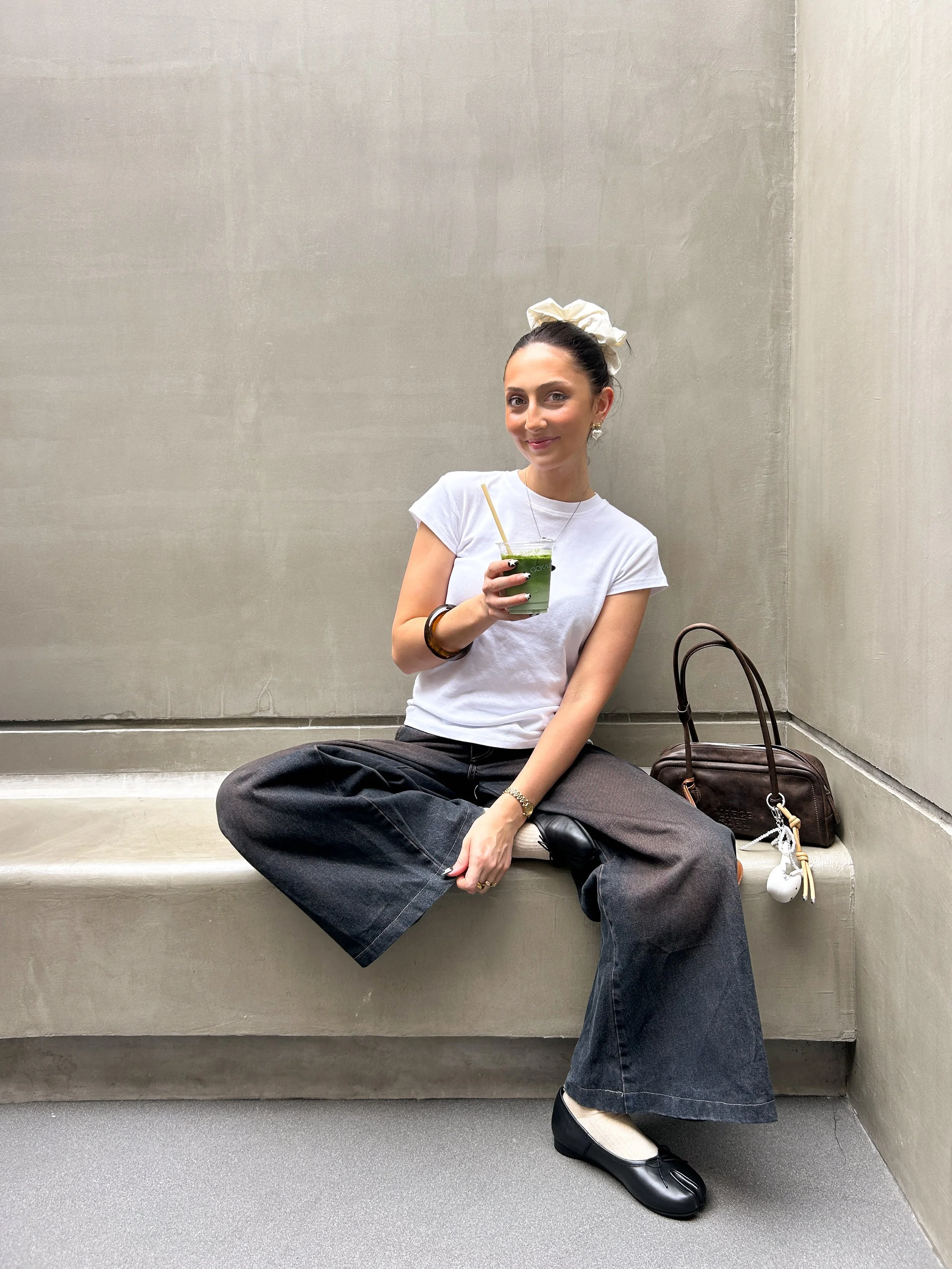 A woman sitting on a concrete bench, wearing a white T-shirt and wide-leg jeans, holding a green drink with a straw, with a brown purse and white earbud case beside her, against a plain gray wall.