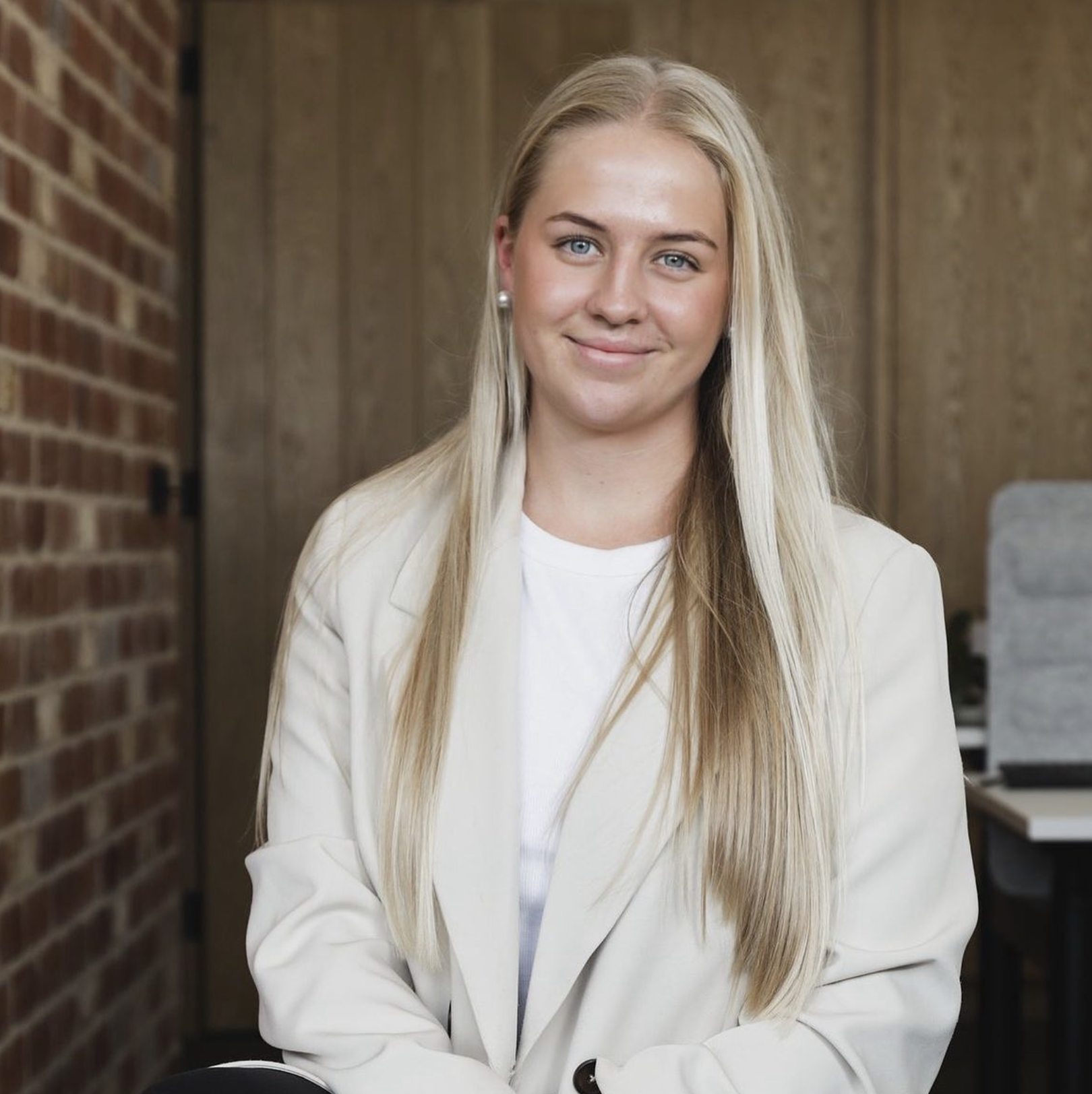 A woman with long blonde hair and blue eyes smiling in an office setting with brick and wood walls.