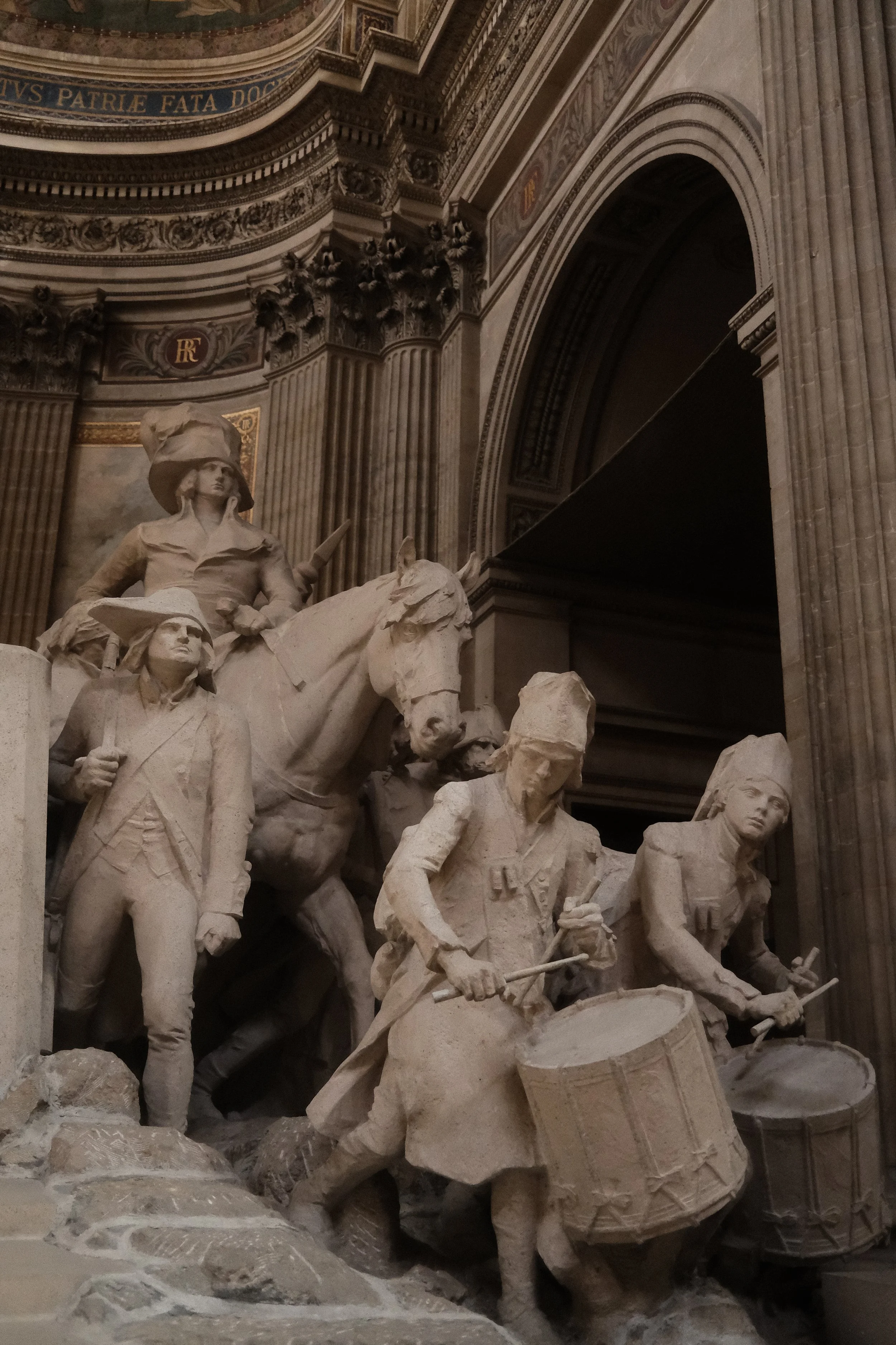 White marble statue of soldiers in combat with guns, drums, and horses inside a grand building with ornate arches and Latin inscriptions.
