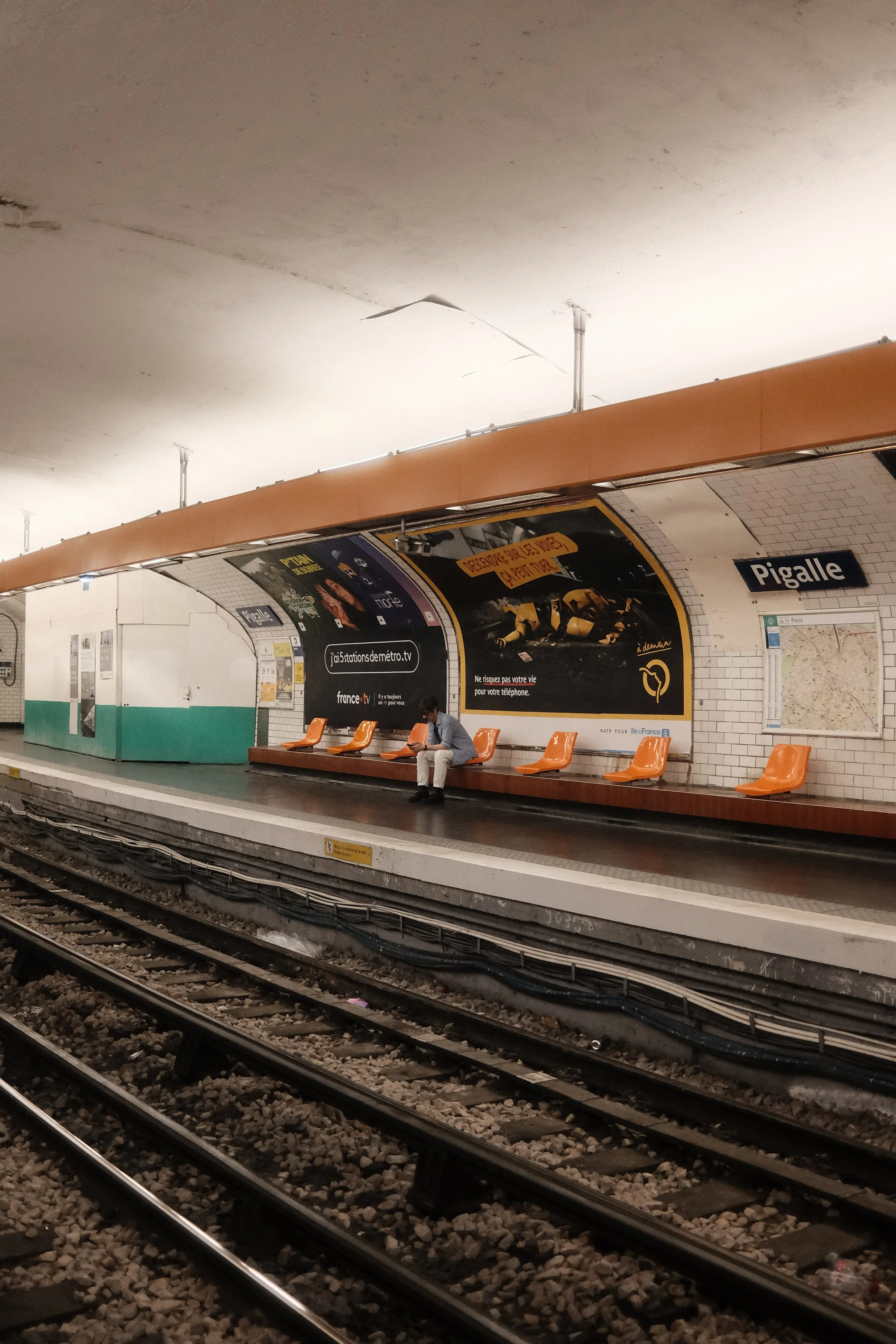 A man sitting on orange benches at Pigalle metro station in Paris, with tracks in the foreground and advertisements on the wall behind.