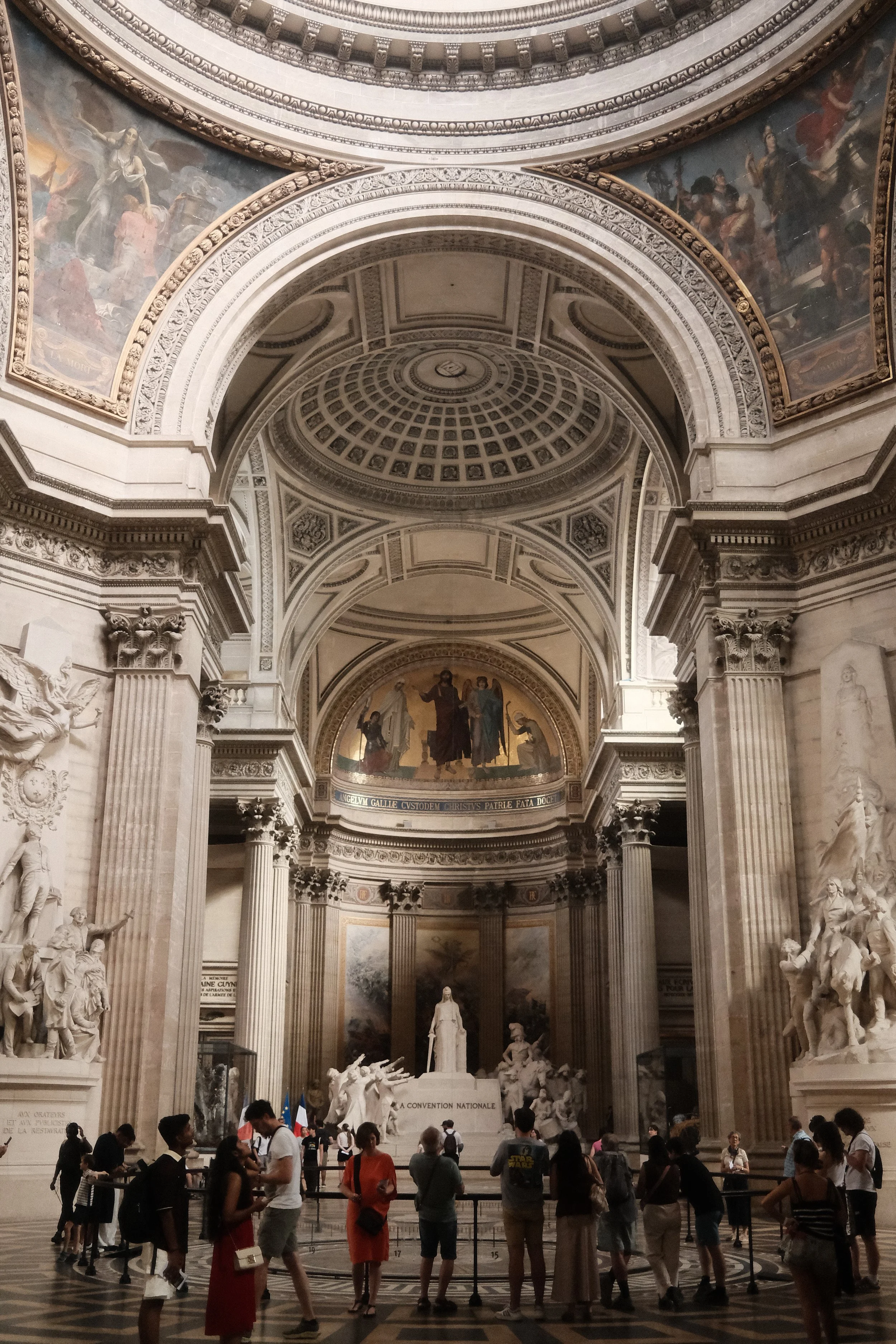 Inside a grand, ornate building with classical architecture, featuring sculptures, columns, and detailed artwork. People are gathered, some looking at displays or statues, in front of a large central statue, with paintings and decorative arches in th