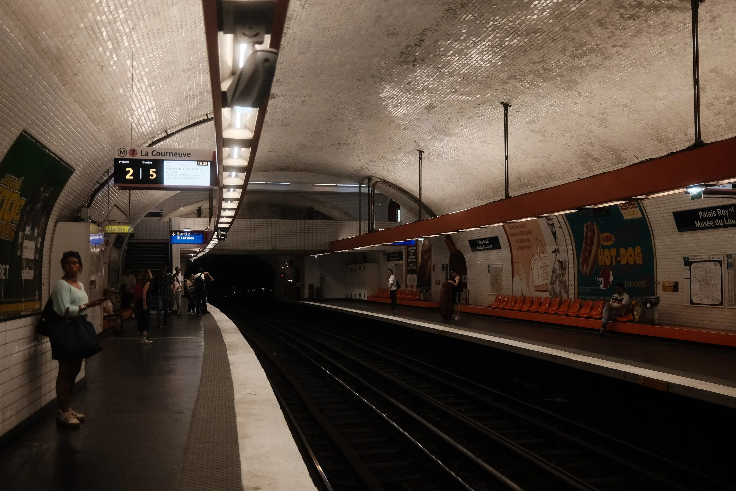 Inside a Paris metro station, several people are waiting on the platform. The curved ceiling is lined with small tiles, and there are signs indicating the station name 'La Courneuve' and directions for exiting and train lines. Orange seating is along