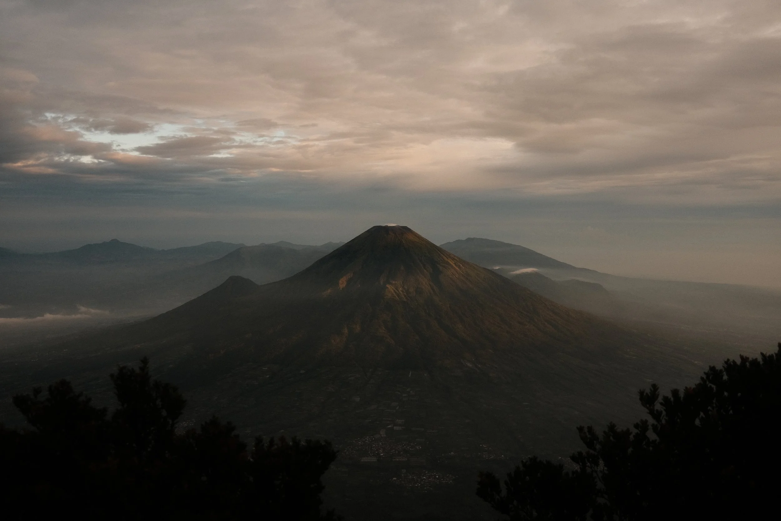 A mountain landscape with a prominent volcano in the center under a cloudy sky during dawn or dusk.