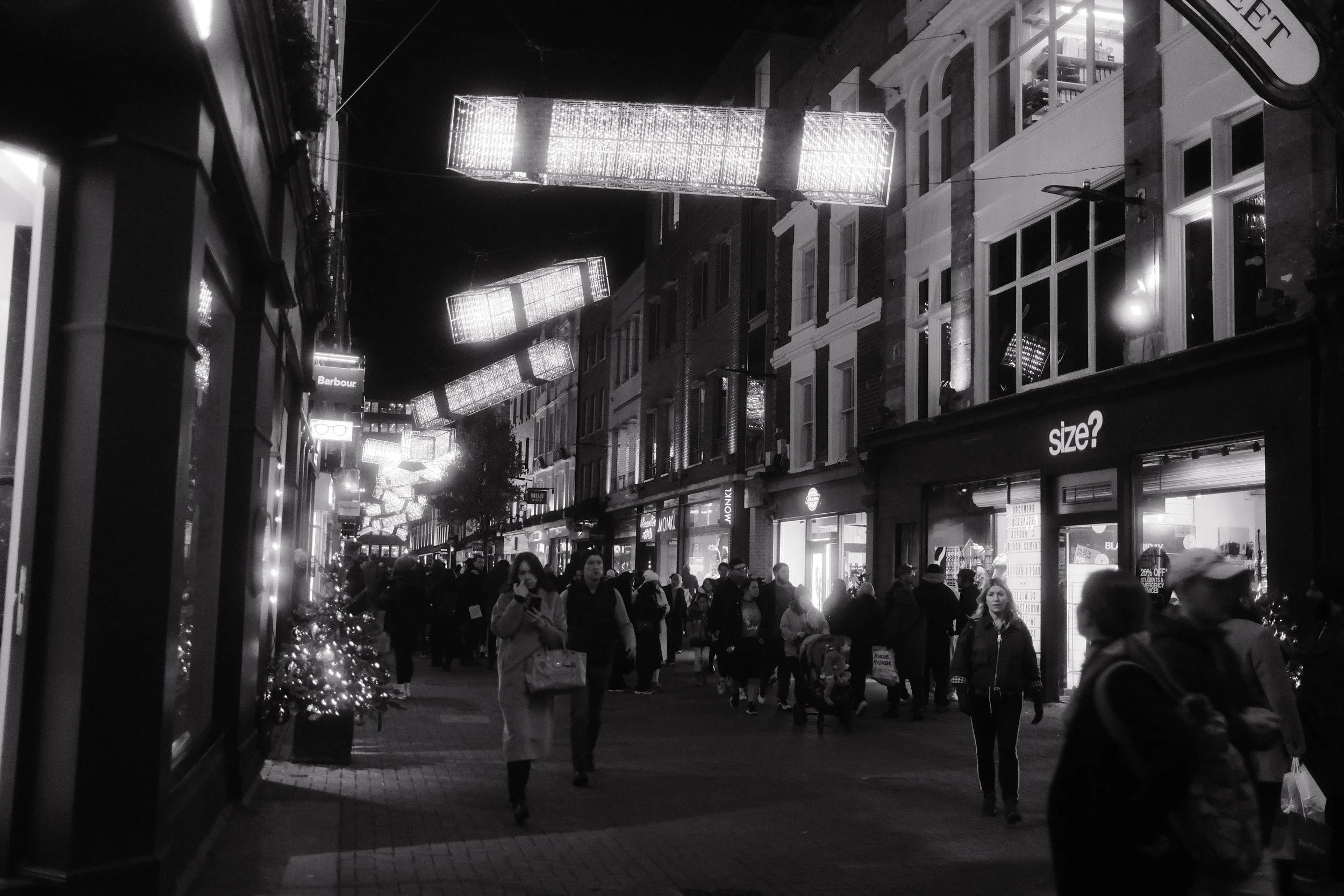 A busy city street at night decorated with hanging illuminated light fixtures. Crowds of people are walking along the sidewalk amidst storefronts and buildings.