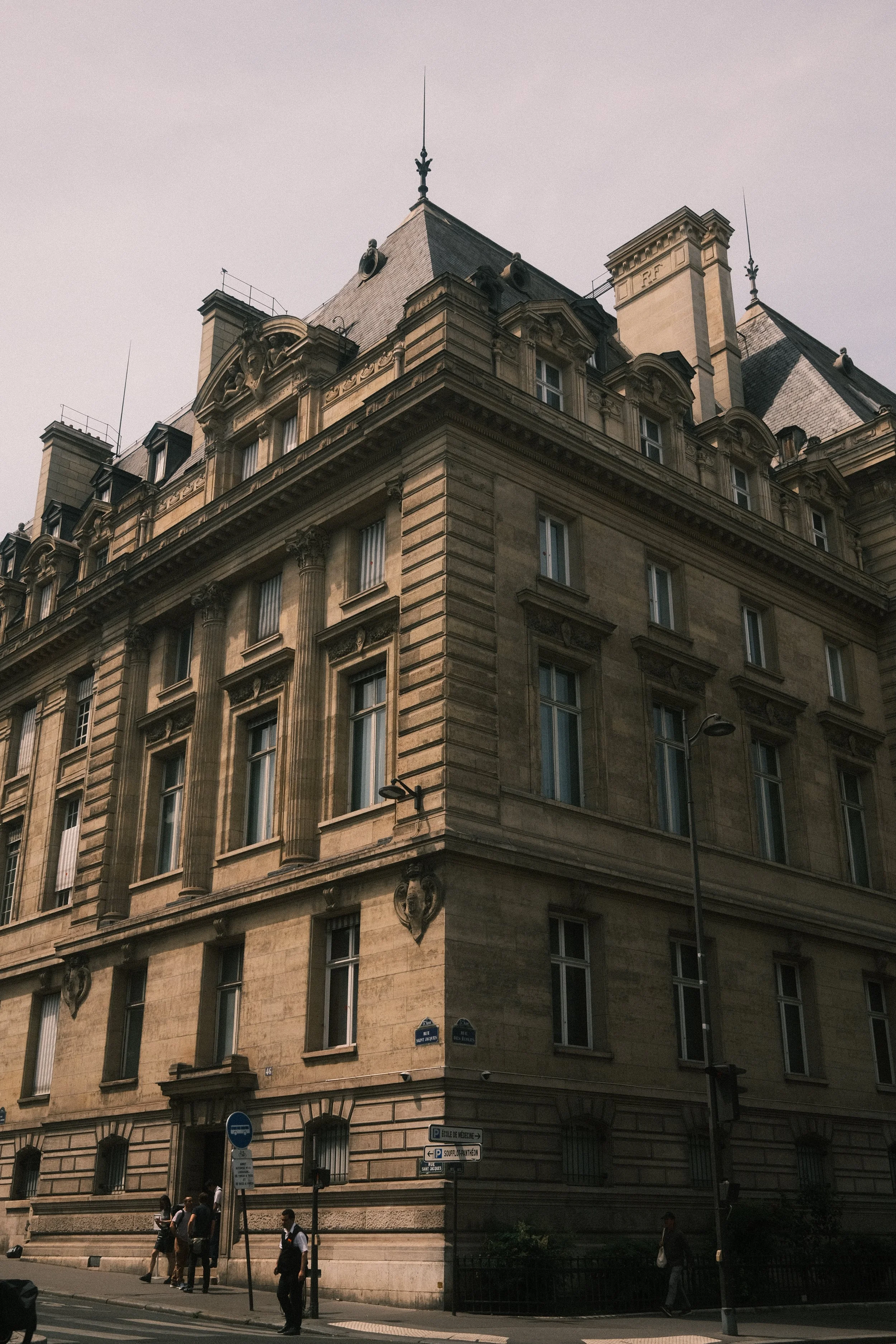 A historic stone building with classical architectural details, including decorative columns and sculptures, and tall mansard roofs with dormer windows. People are walking on the sidewalk in front of the building.