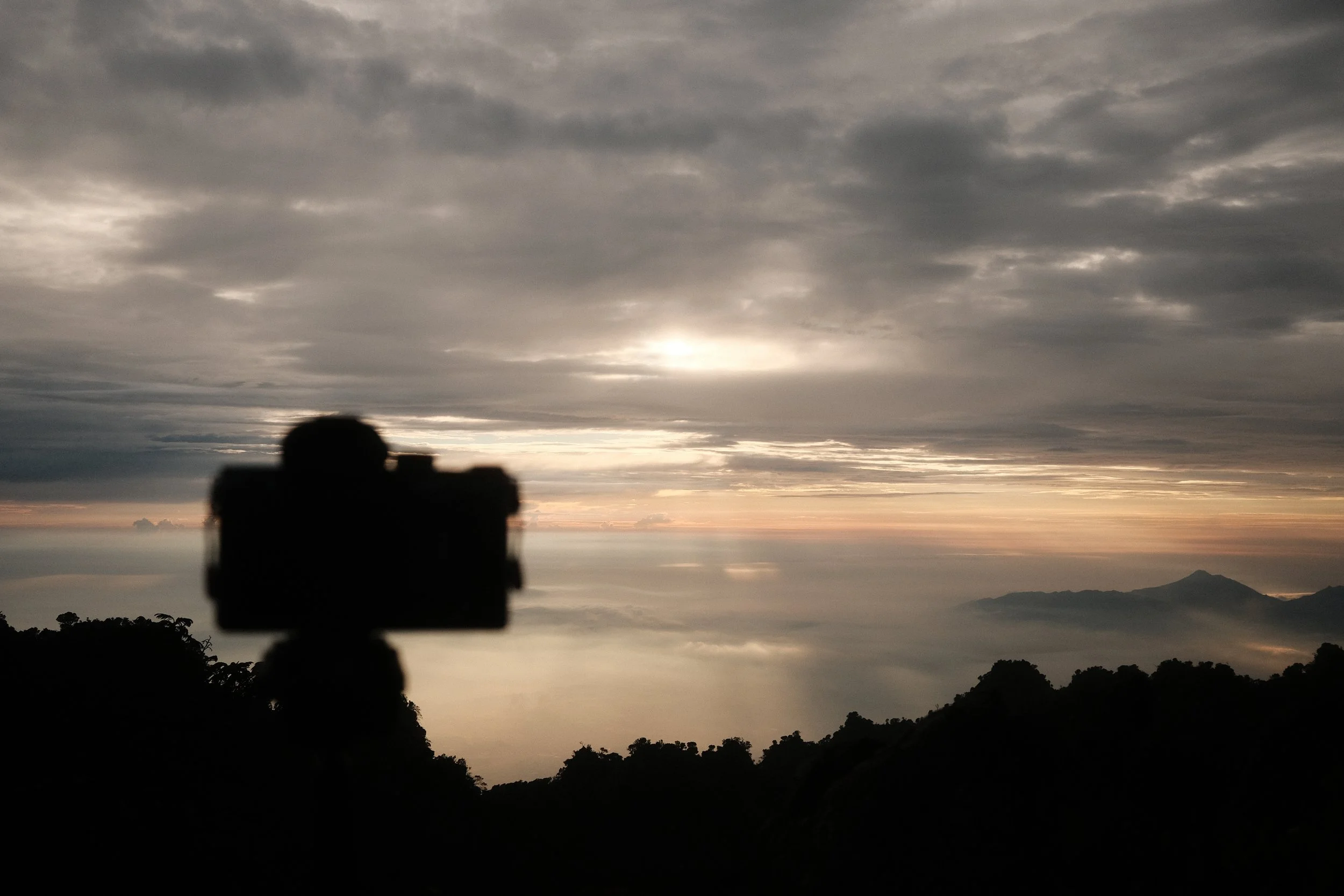 A landscape scene of a cloudy sky during sunset or sunrise with a silhouette of mountains and trees in the foreground, and a blurry object that appears to be a camera or binoculars in the bottom center.