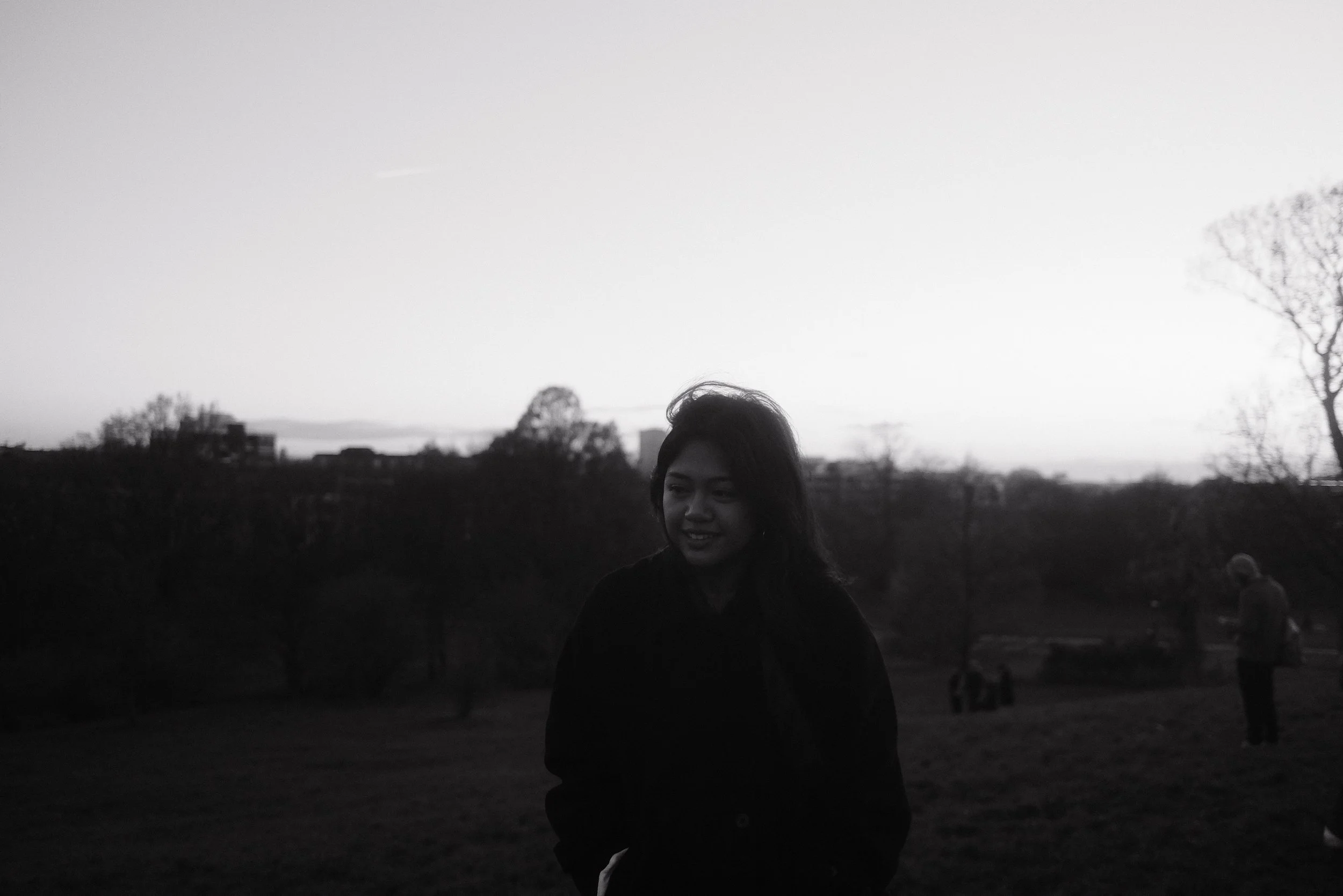 A black-and-white photo of a woman outdoors in a park at sunset or dusk, with trees and a skyline in the background.