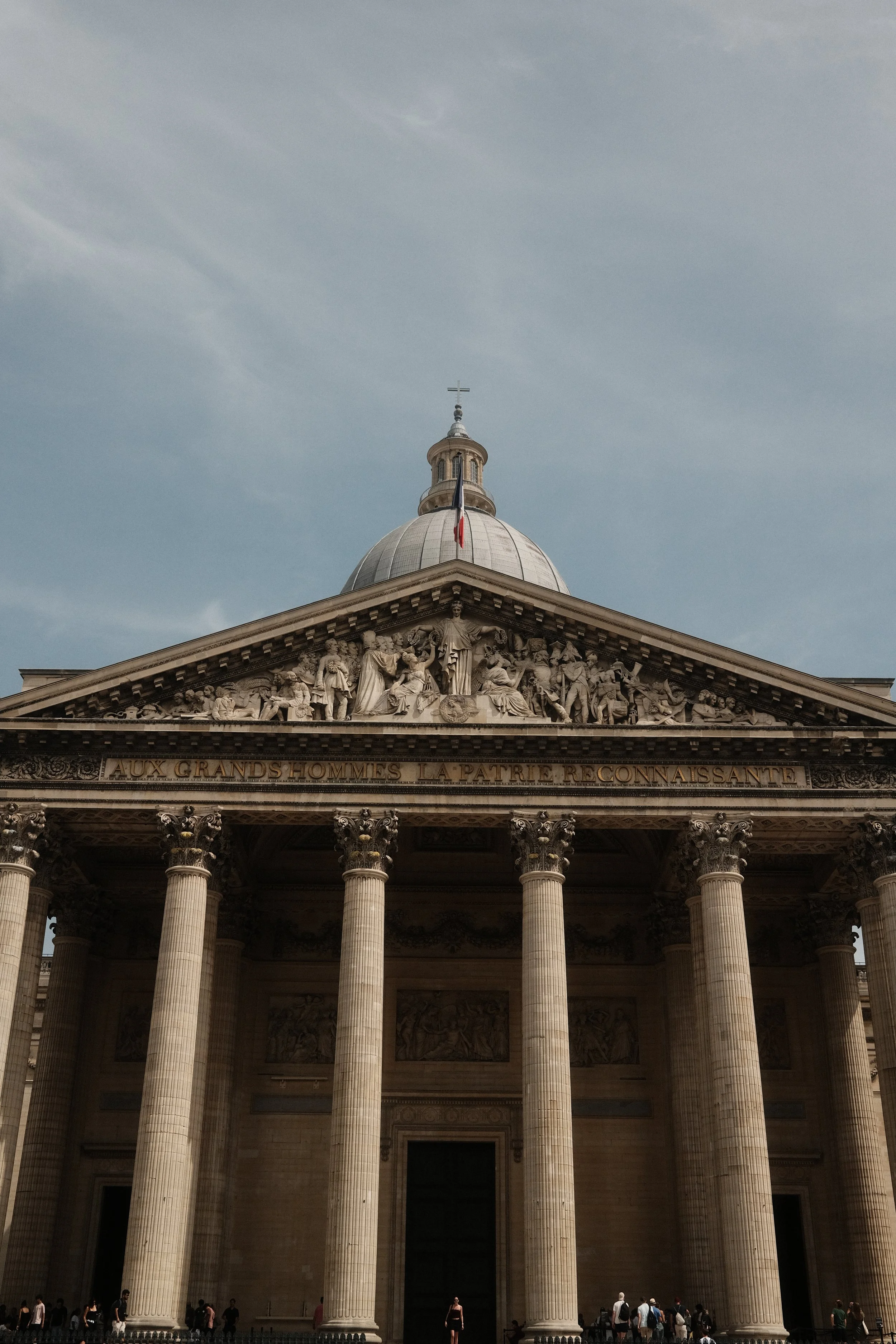 The facade of a classical building with tall columns, a detailed pediment with sculptures, and a dome topped with a cross. There are people walking in front of the building.