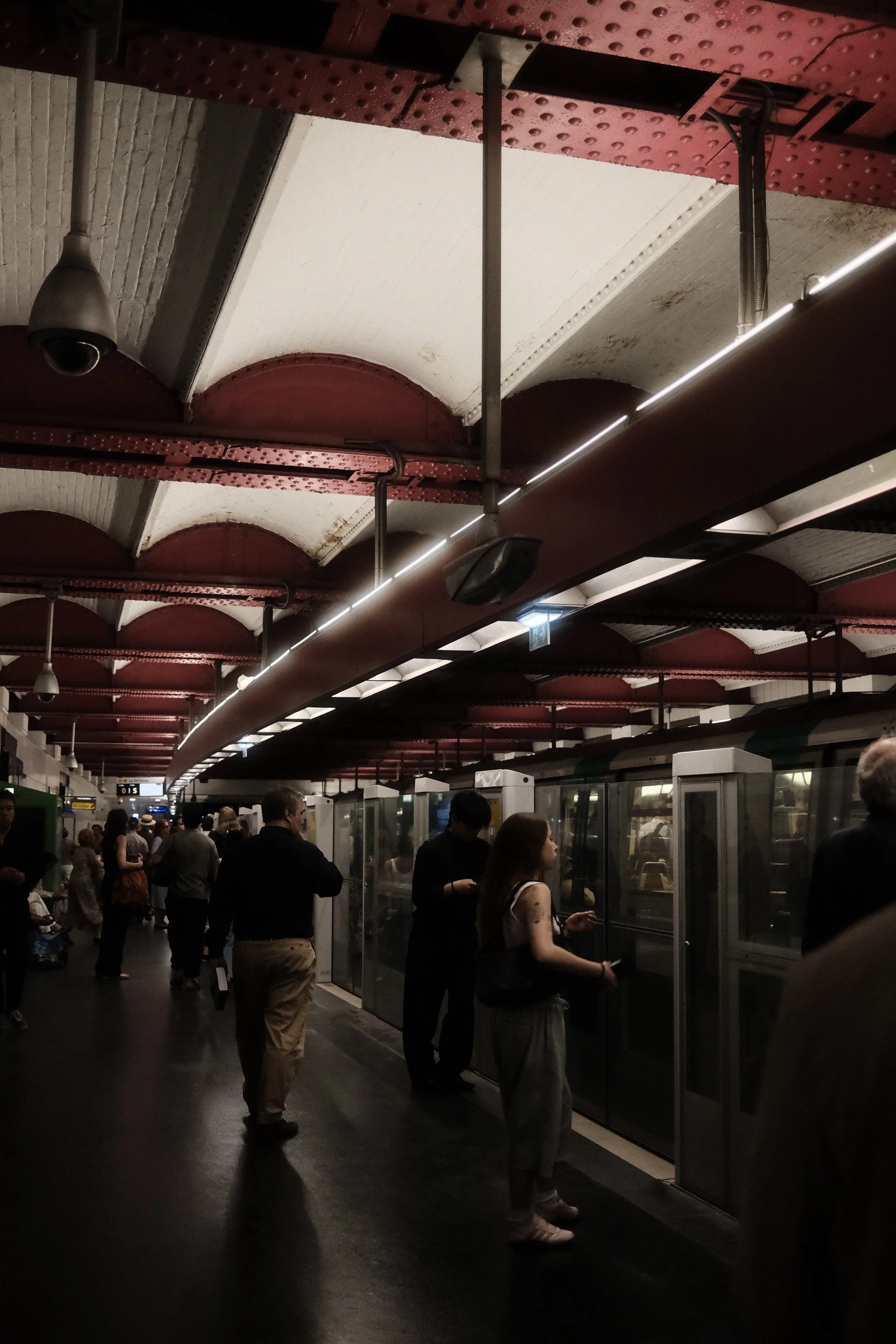 People waiting in line at a train station with glass doors, crowds under a decorated arched ceiling with red and white patterns, and surveillance cameras visible.