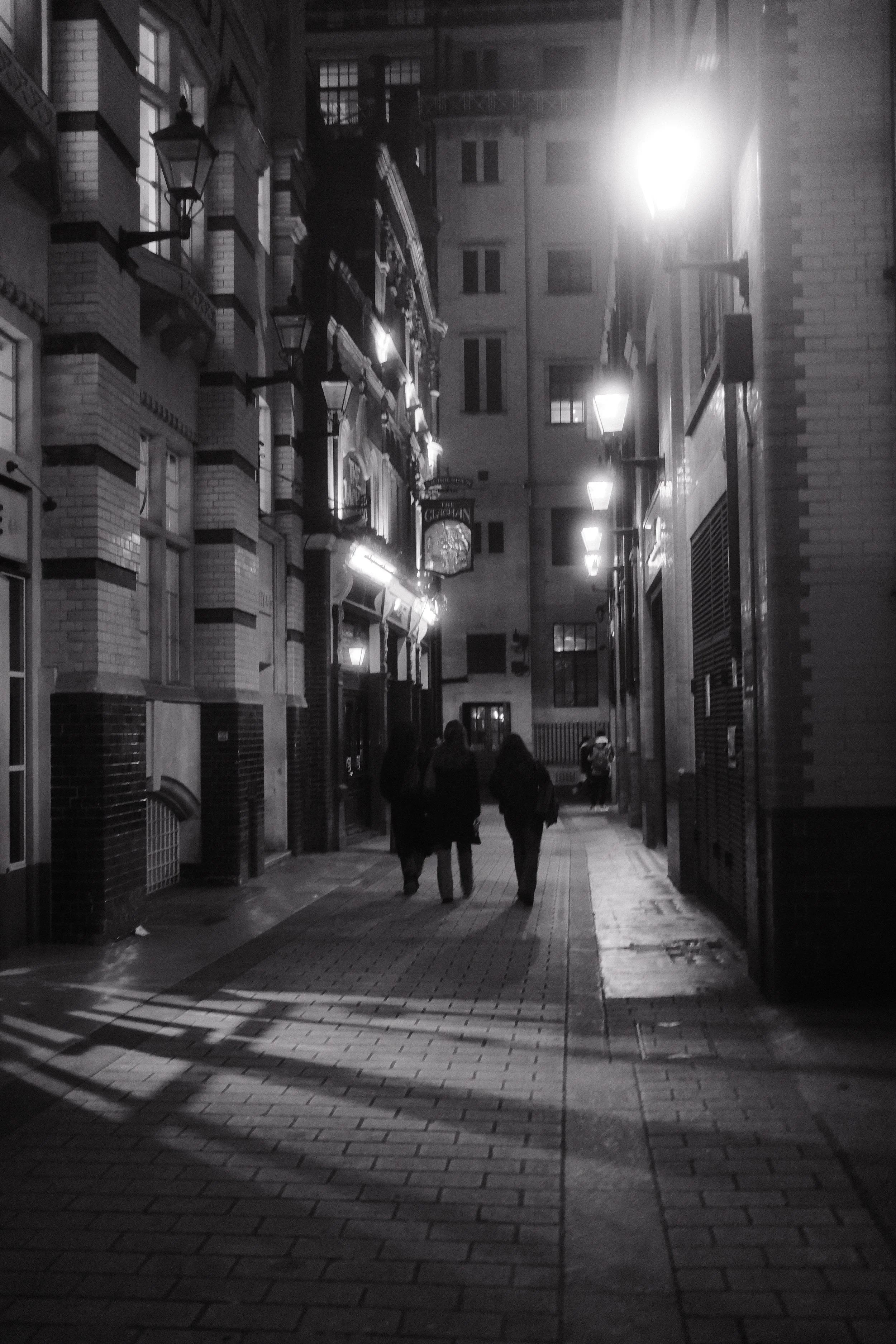 A black and white night street scene with a narrow alley lined with buildings. Street lamps illuminate the area, and a few people are walking along the sidewalk.