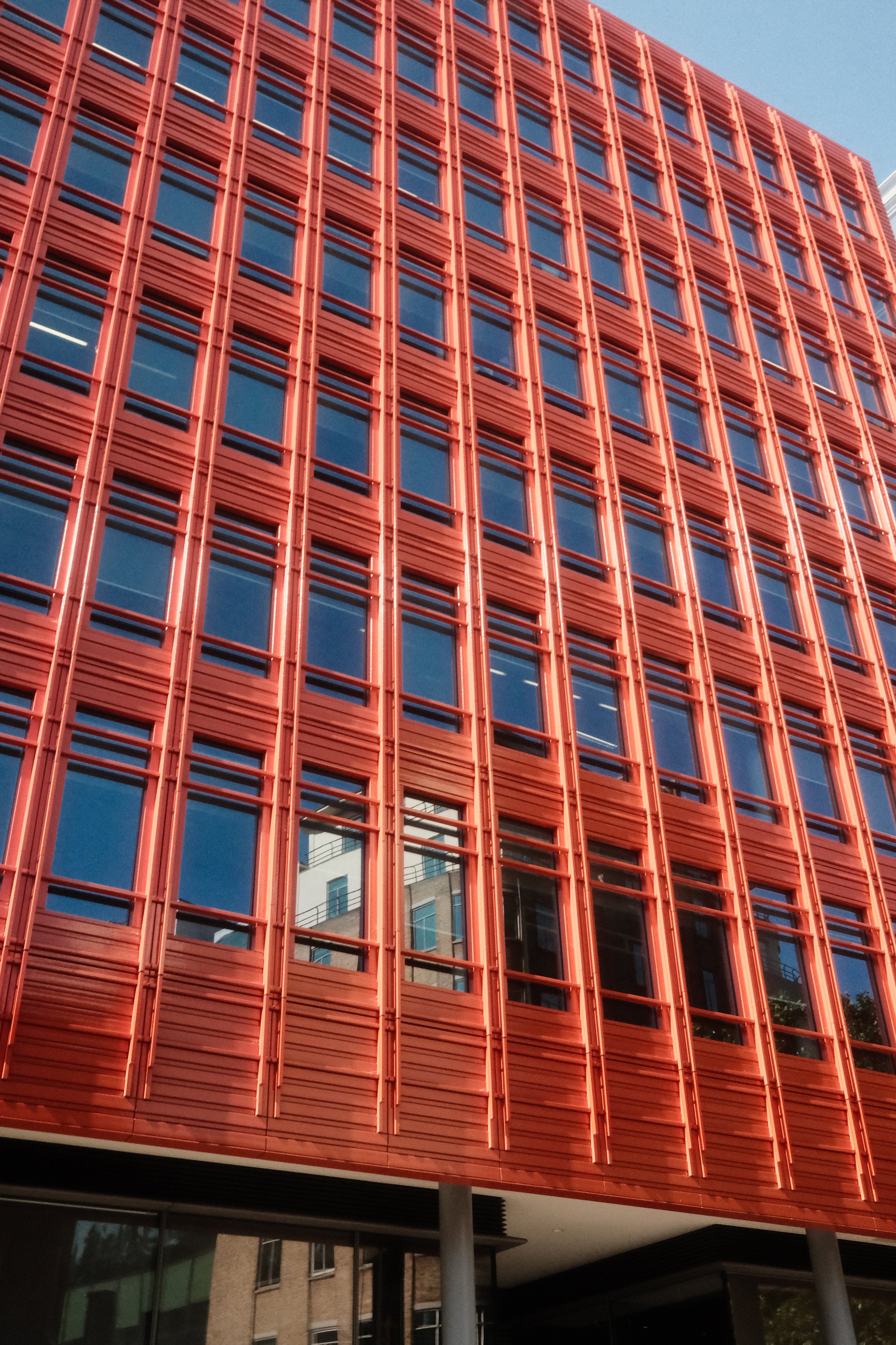 Close-up of a modern building exterior with red metallic grid-like paneling and reflective glass windows, with some portions of the building and surrounding structures visible below.