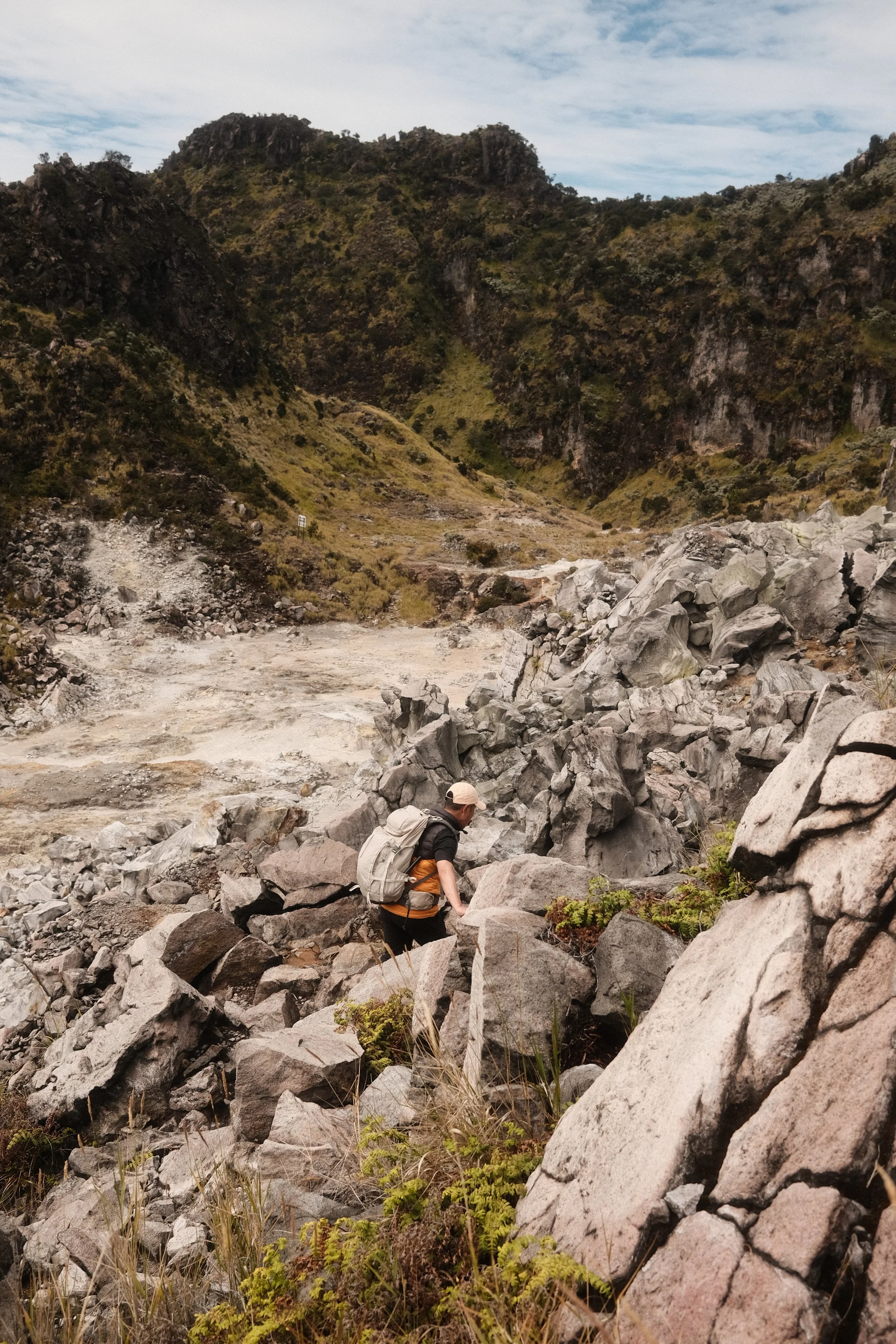 A person with a backpack hiking over large rocks in a rugged, mountain landscape with steep cliffs and sparse vegetation under a cloudy sky.