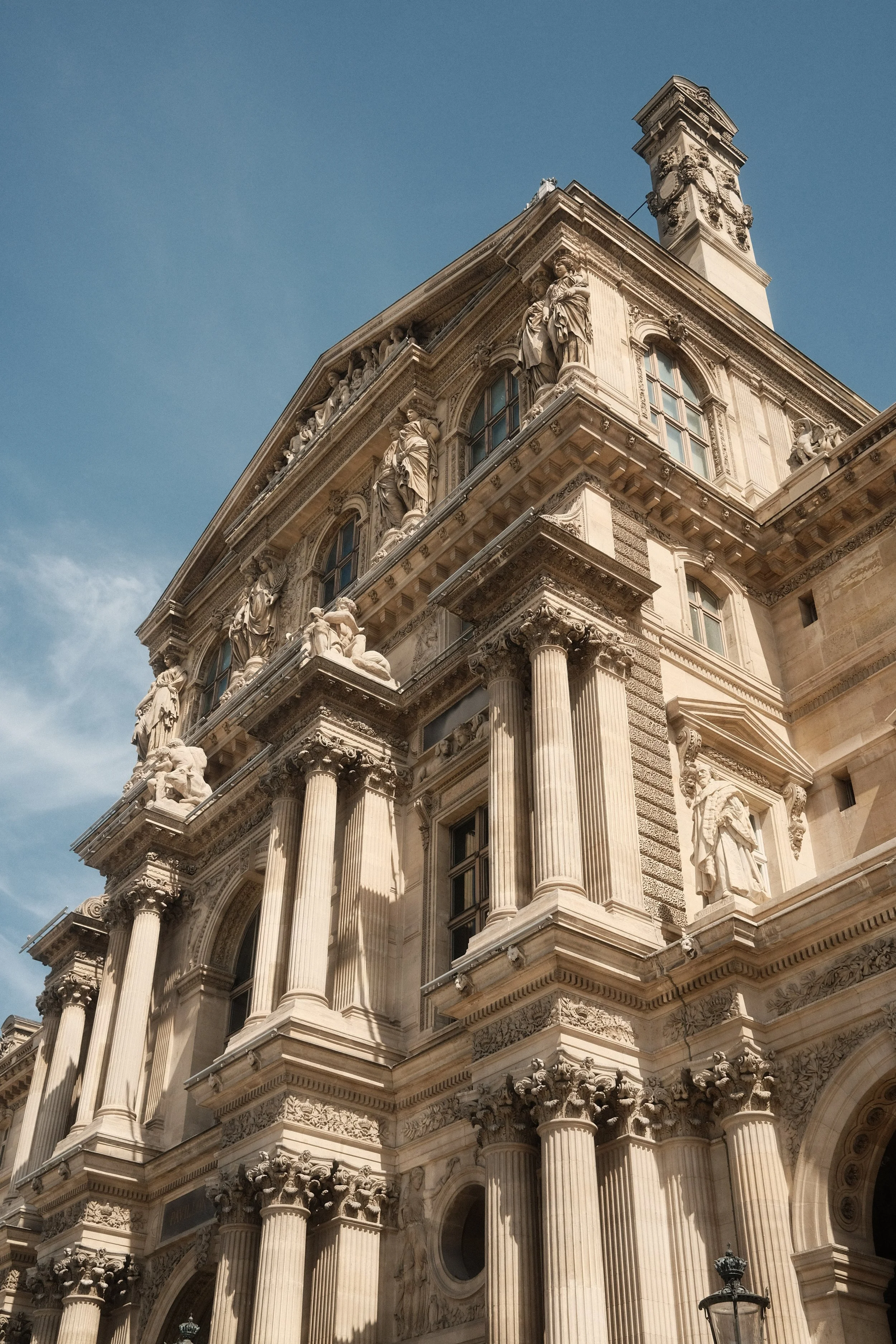 The ornate facades of a historic building with classical architecture, including Corinthian columns, statues, and decorative sculptures, under a blue sky.