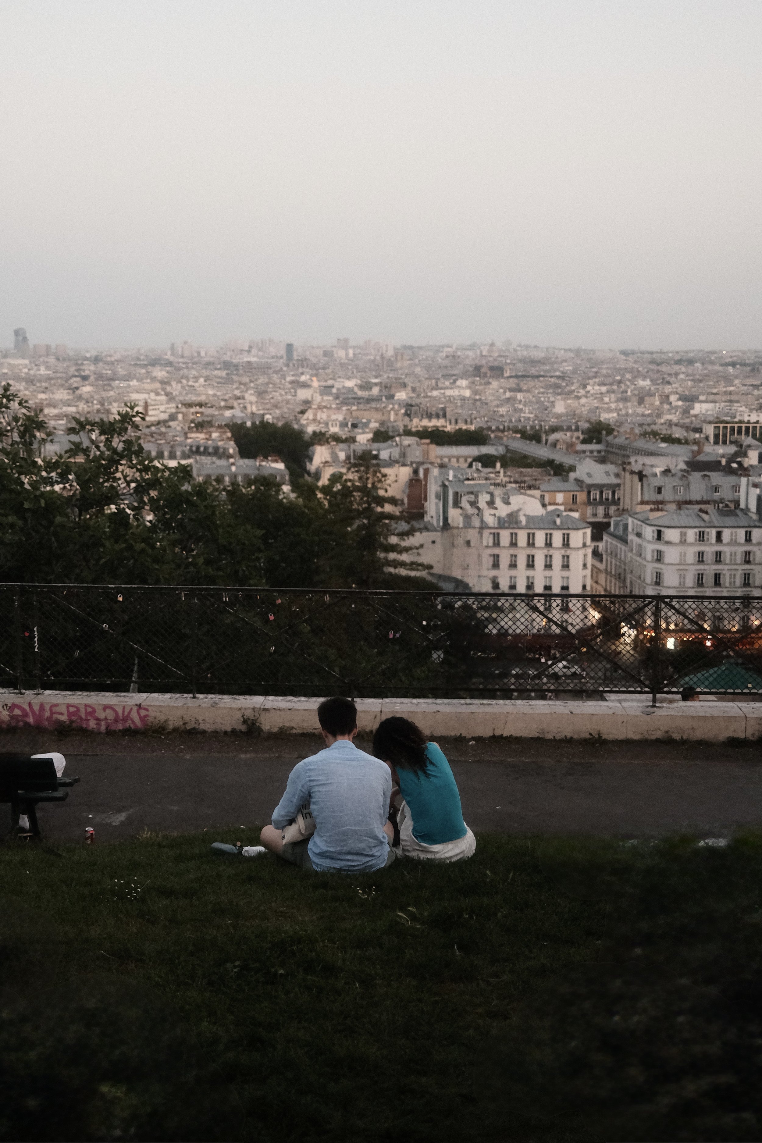 Two people sitting on grass near a fence overlooking a sprawling cityscape of Paris with many buildings and trees during dusk.