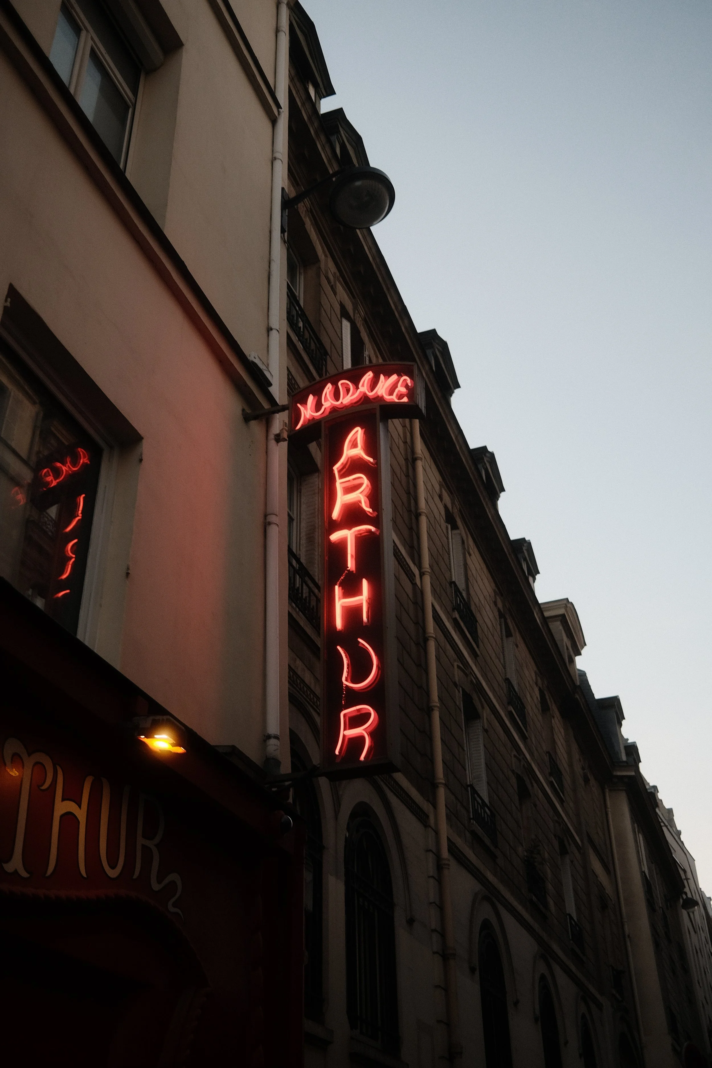 A vertical neon sign on the side of a building reads 'HOTEL'. The sign is lit in red against a darkening sky, with a street lamp and the upper floors of the building also visible.