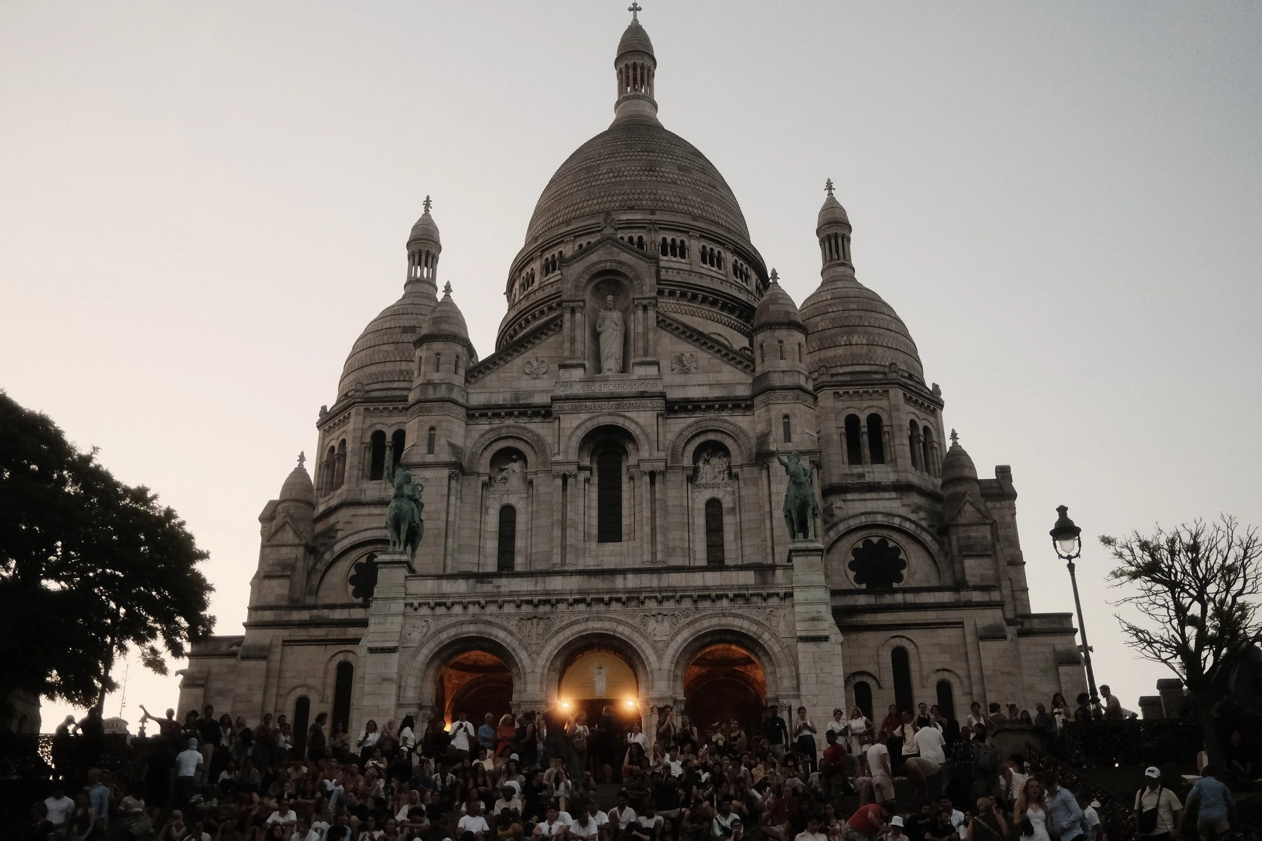 View of the Sacré-Cœur Basilica in Paris, France, with a large crowd of people gathered on the steps and around the entrance during dusk, with the sky turning gray and several trees in the foreground.