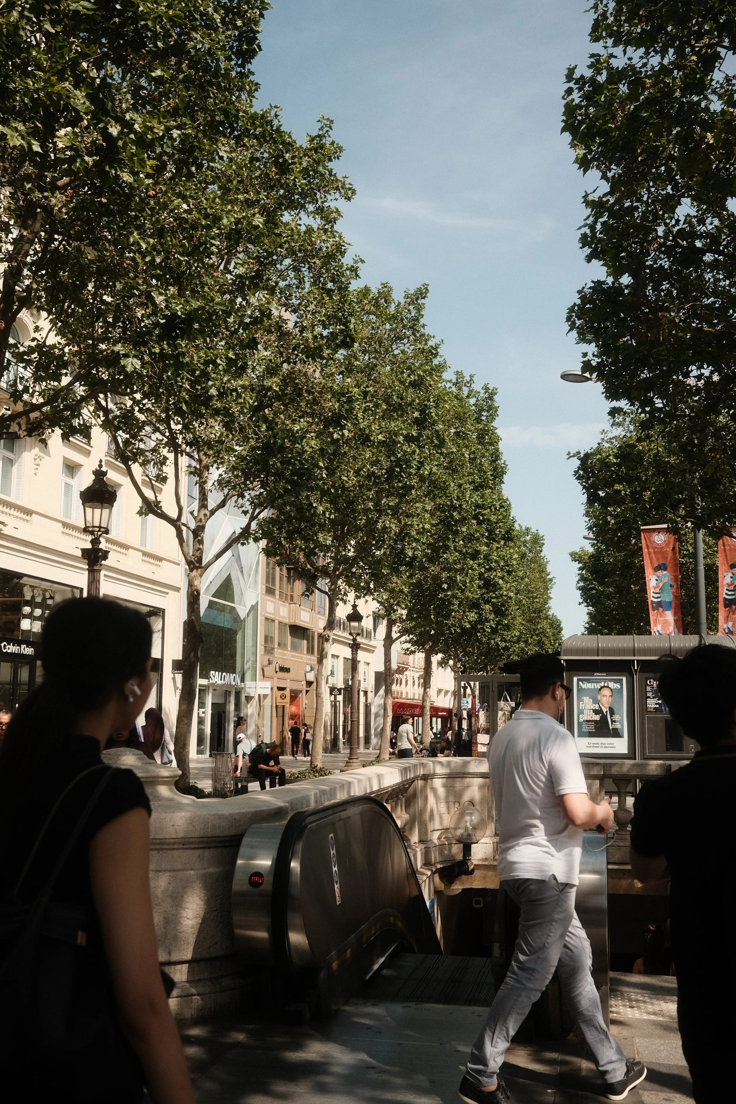 Urban street scene with people walking, trees lining the sidewalk, storefronts including Calvin Klein, Salomon, and other shops, and advertisements in the background.