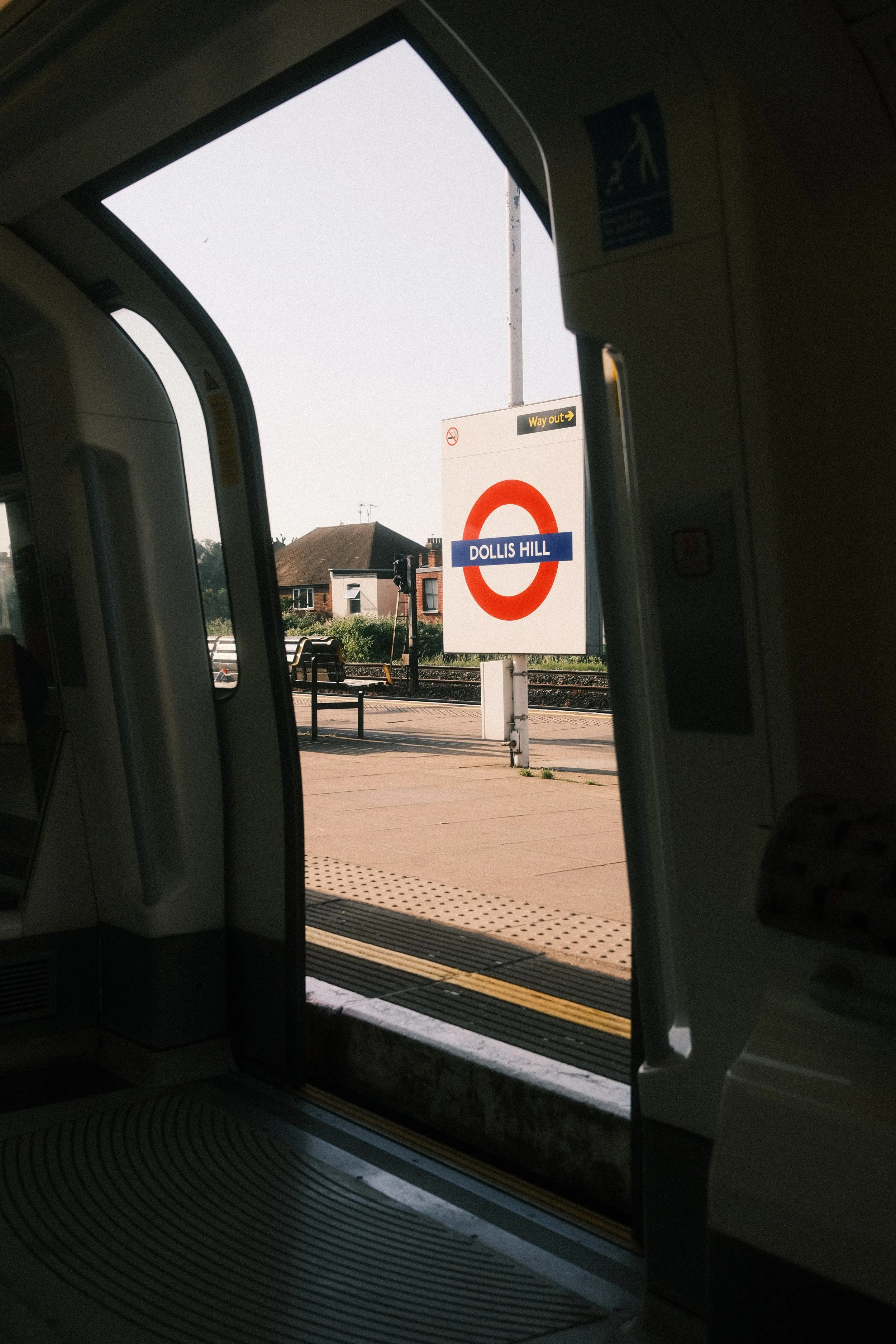 View from inside a train looking out onto the platform at Dollis Hill station, with a sign indicating the station name and an exit sign visible in the background.