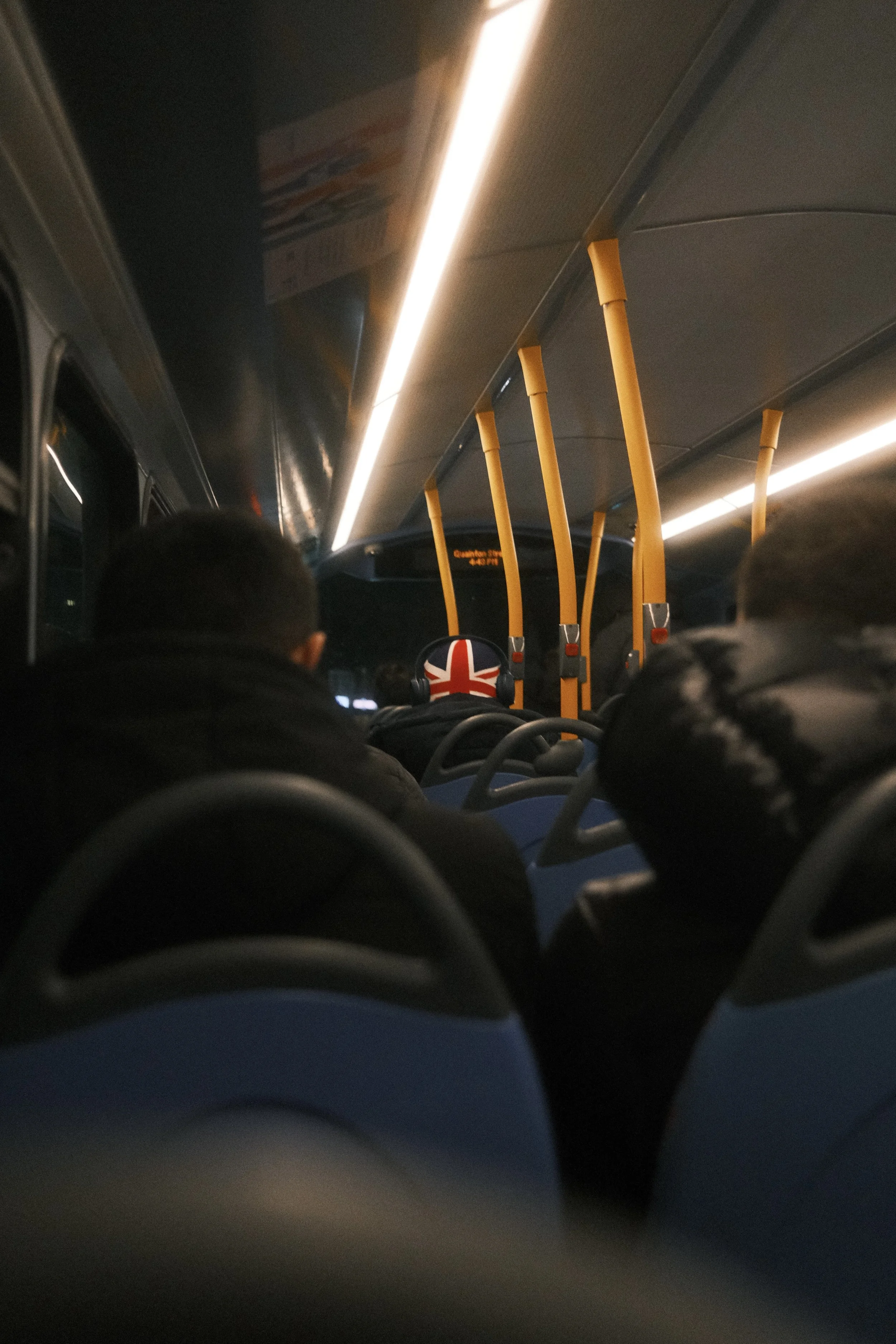 Inside a bus during nighttime, showing the backs of several passengers seated and the yellow poles and handrails, with a digital display at the front showing the destination.
