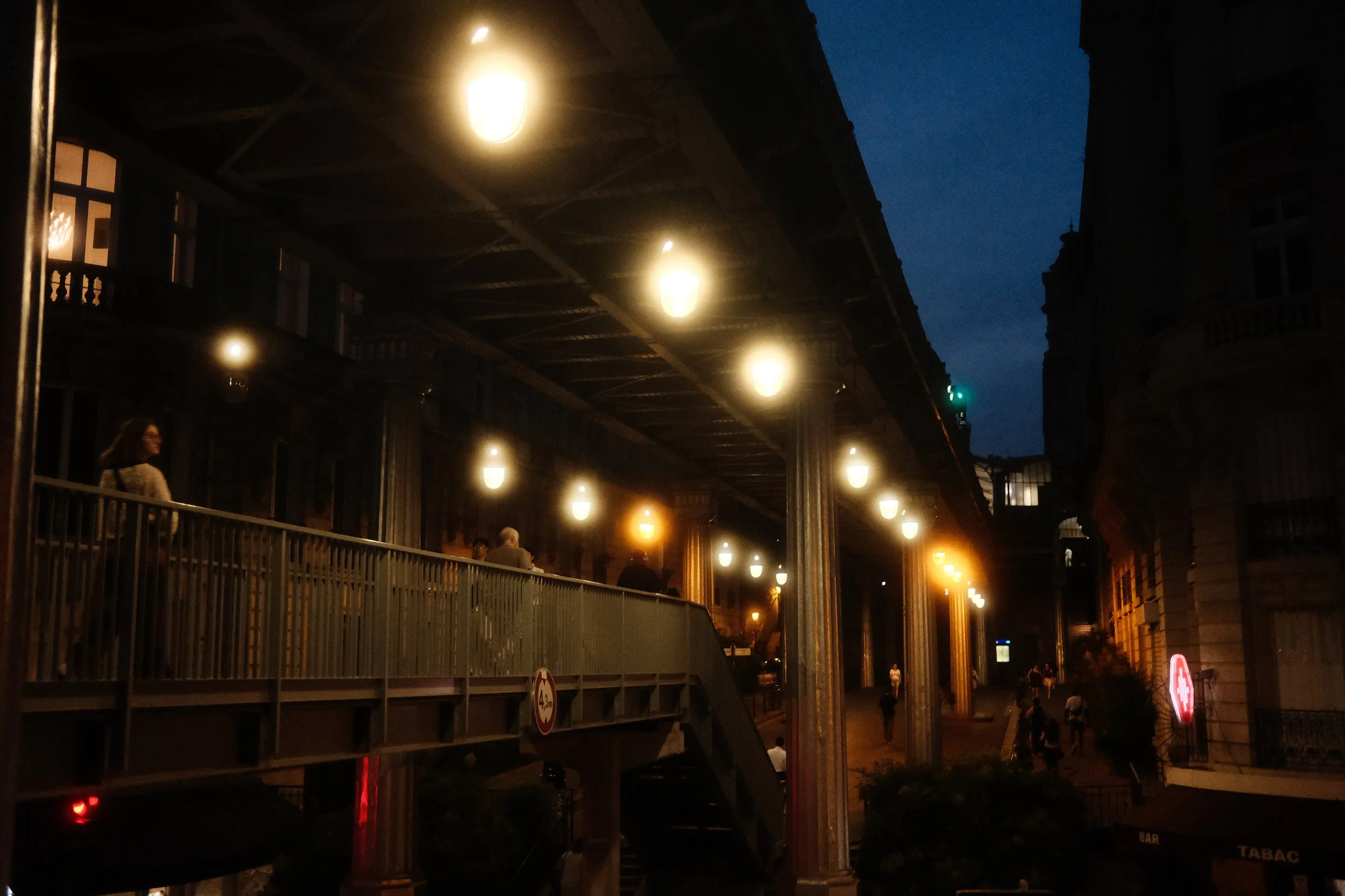 Nighttime scene of a city street with a elevated train station and hanging lights, people walking below and some standing on the station walkway.