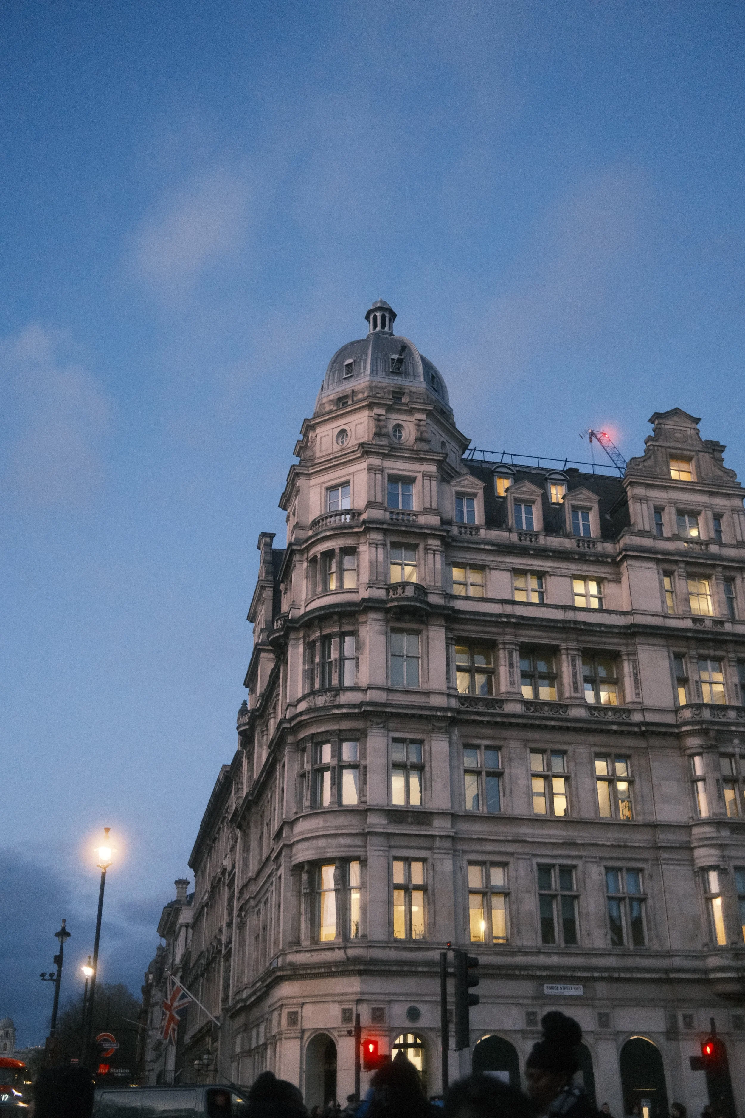A tall, historic building with a rounded top and multiple windows, set against a dusky evening sky. There are streetlights, pedestrians, a traffic light, and Union Jack flags visible in the scene.