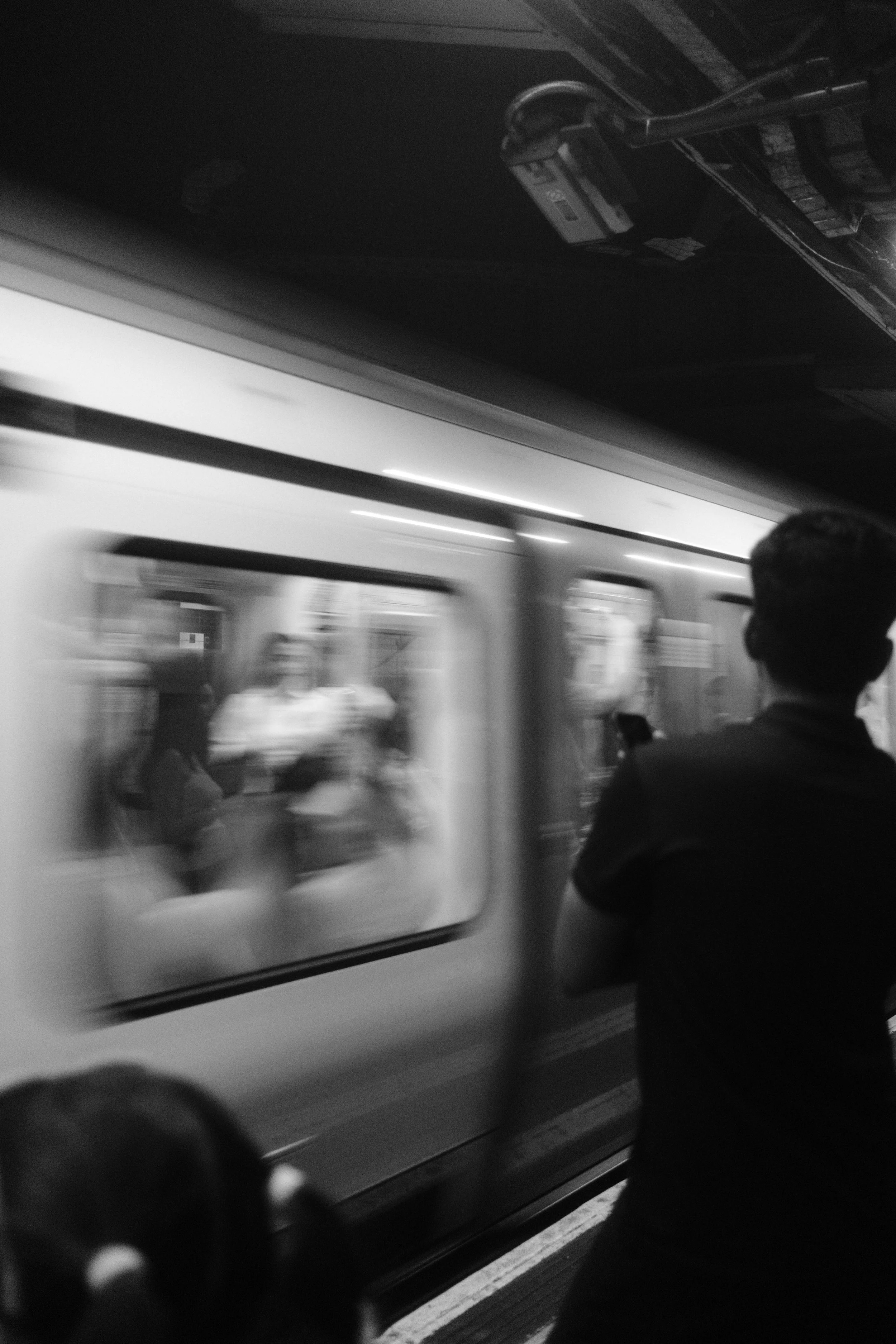 A train is arriving at a station, with a person standing on the platform looking at their phone. The train has a large advertisement on its side.