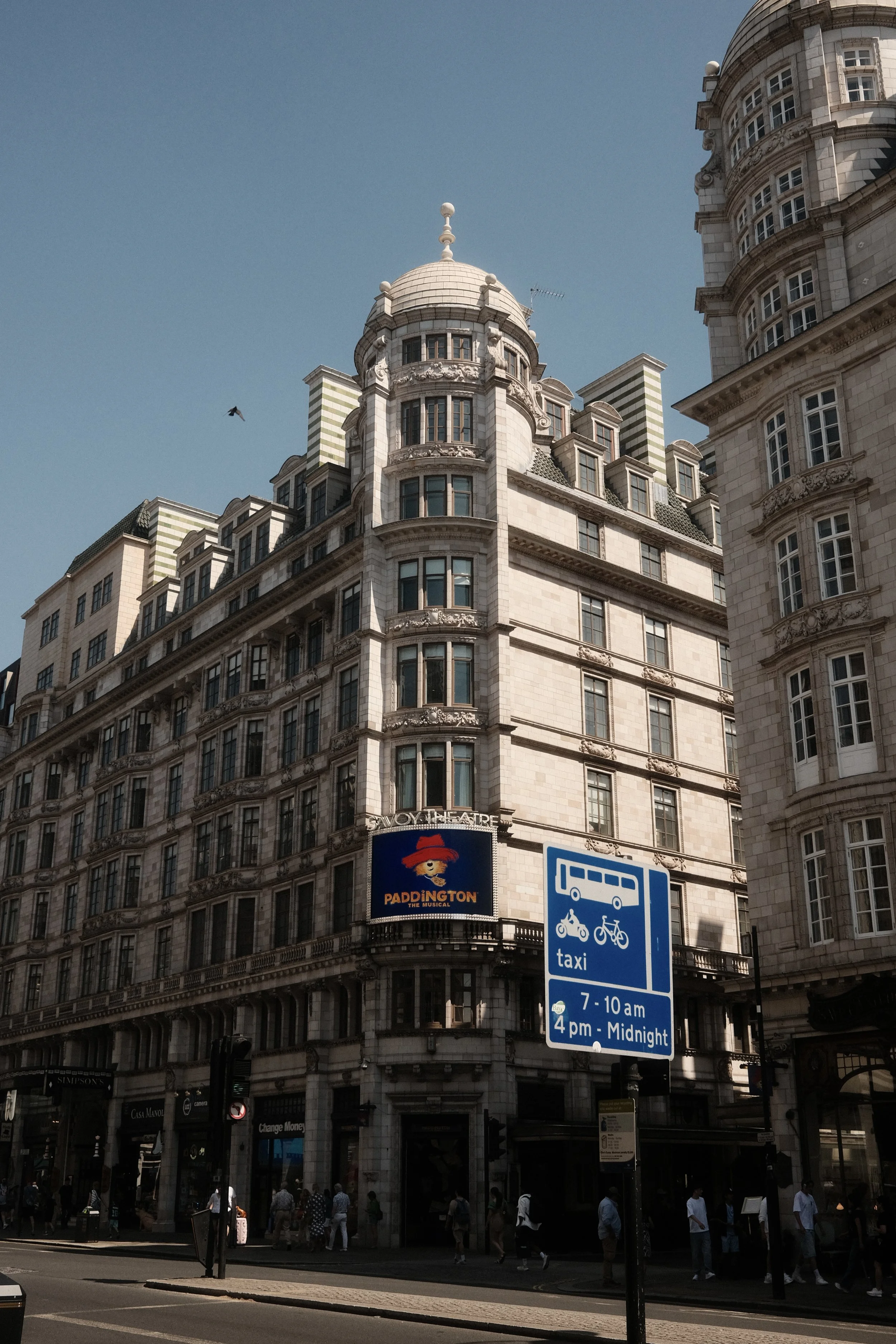 The image shows the upper part of a historic building with ornate architecture on a city street. There is a sign advertising 'Paddington the Musical' hanging from the building. Pedestrians are walking on the sidewalk, and a blue sign indicating bus, 