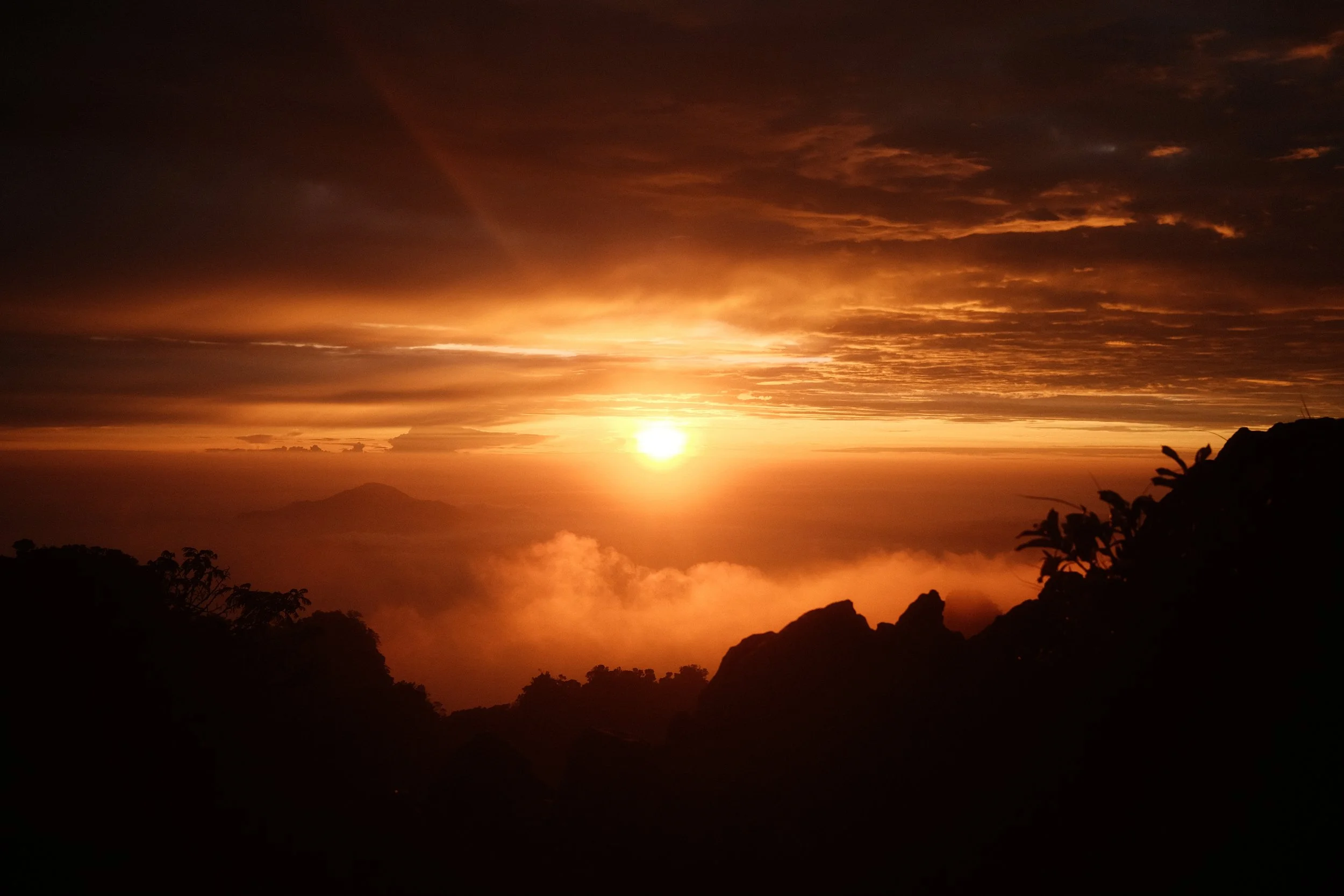 Sunset over mountains with a colorful sky and clouds, silhouetted trees and rocks in the foreground.