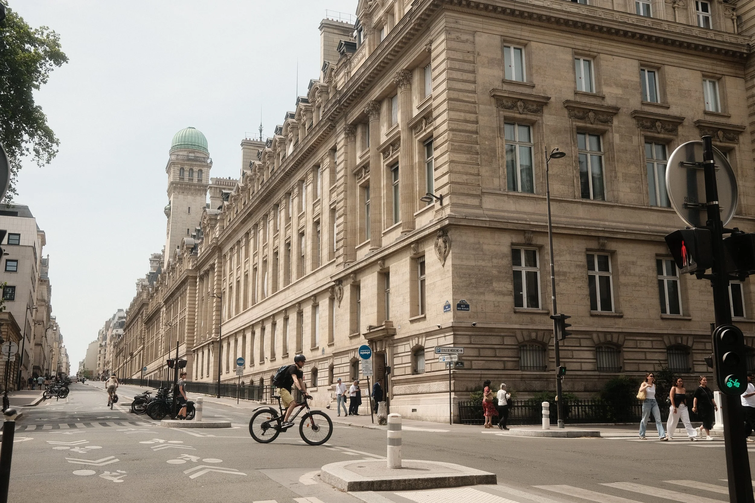 Street scene in a European city with historic beige stone buildings, pedestrians, cyclists, and traffic lights under an overcast sky.