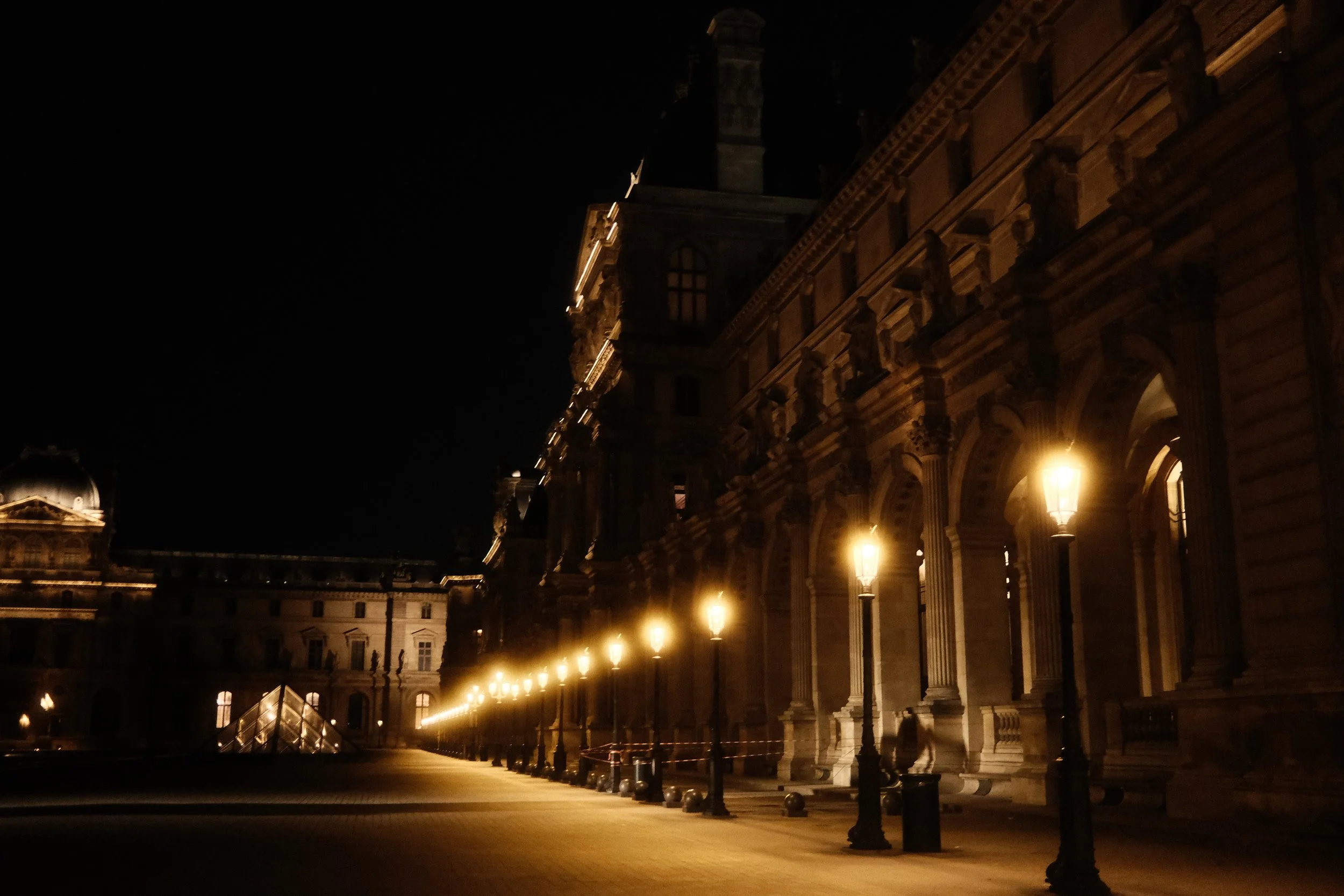 A nighttime view of historic architecture with lit street lamps along the building's exterior, featuring detailed stonework and large arched windows.