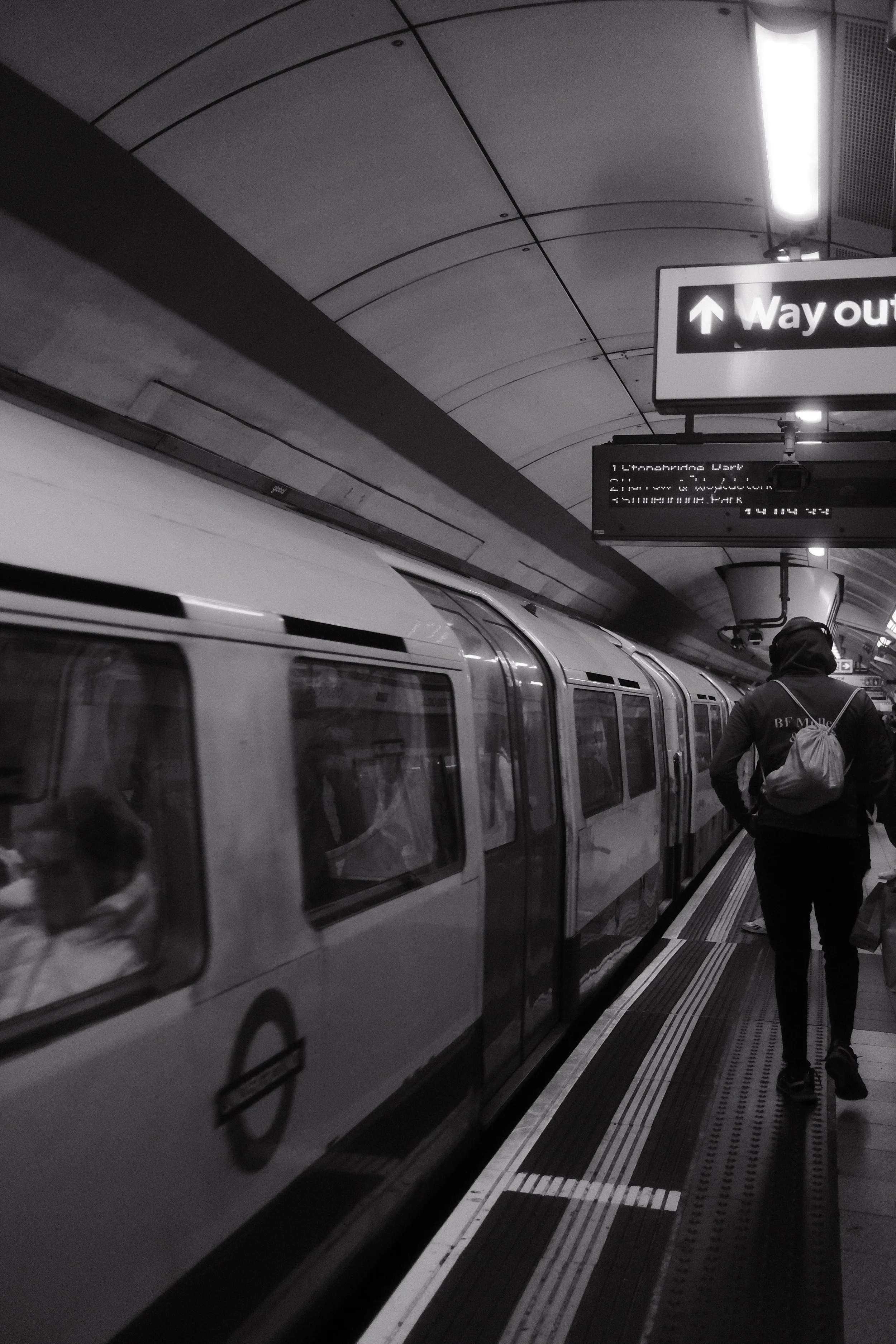 A subway train at an underground station with a person walking on the platform. There are signs indicating the way out and a monitor displaying station information.