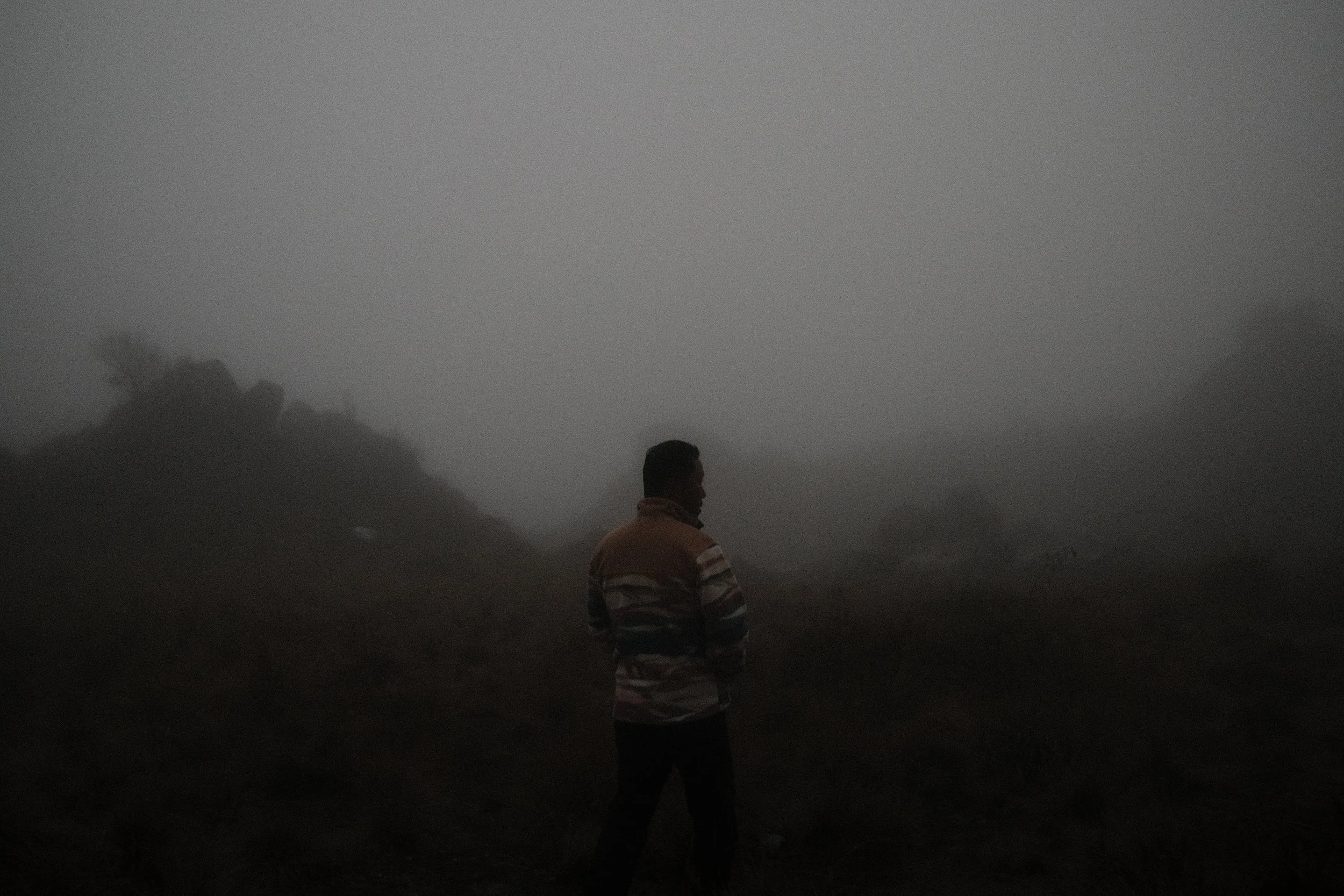 A person standing outdoors on a foggy day, wearing a striped hoodie, with a hillside and trees in the background.