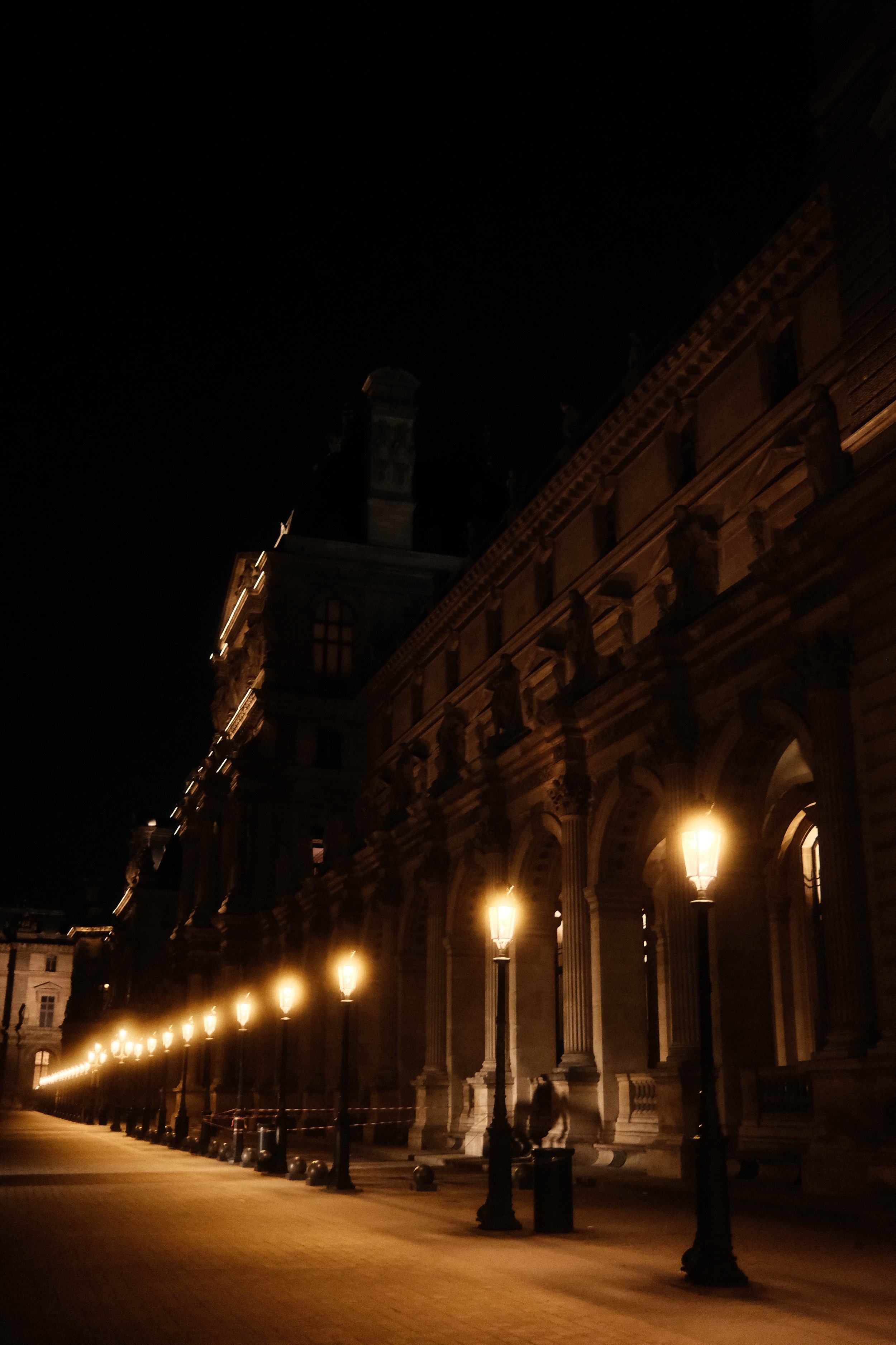 A nighttime view of a historic building with ornate architecture, illuminated by street lamps along a walkway, with no visible people and a dark sky.