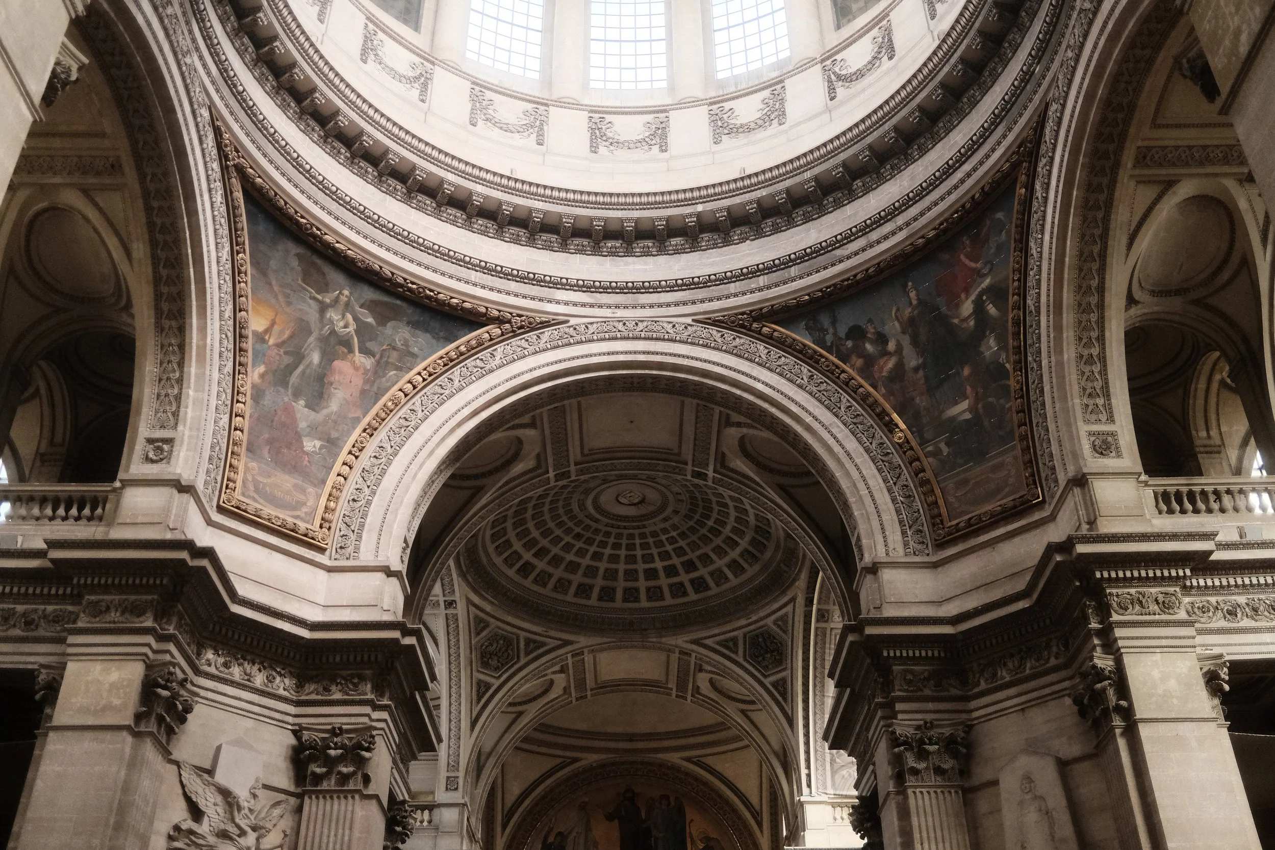The interior of a grand, historic building with ornate architectural details, high arched ceilings, and paintings on the upper walls. The ceiling is illuminated by large windows at the top.