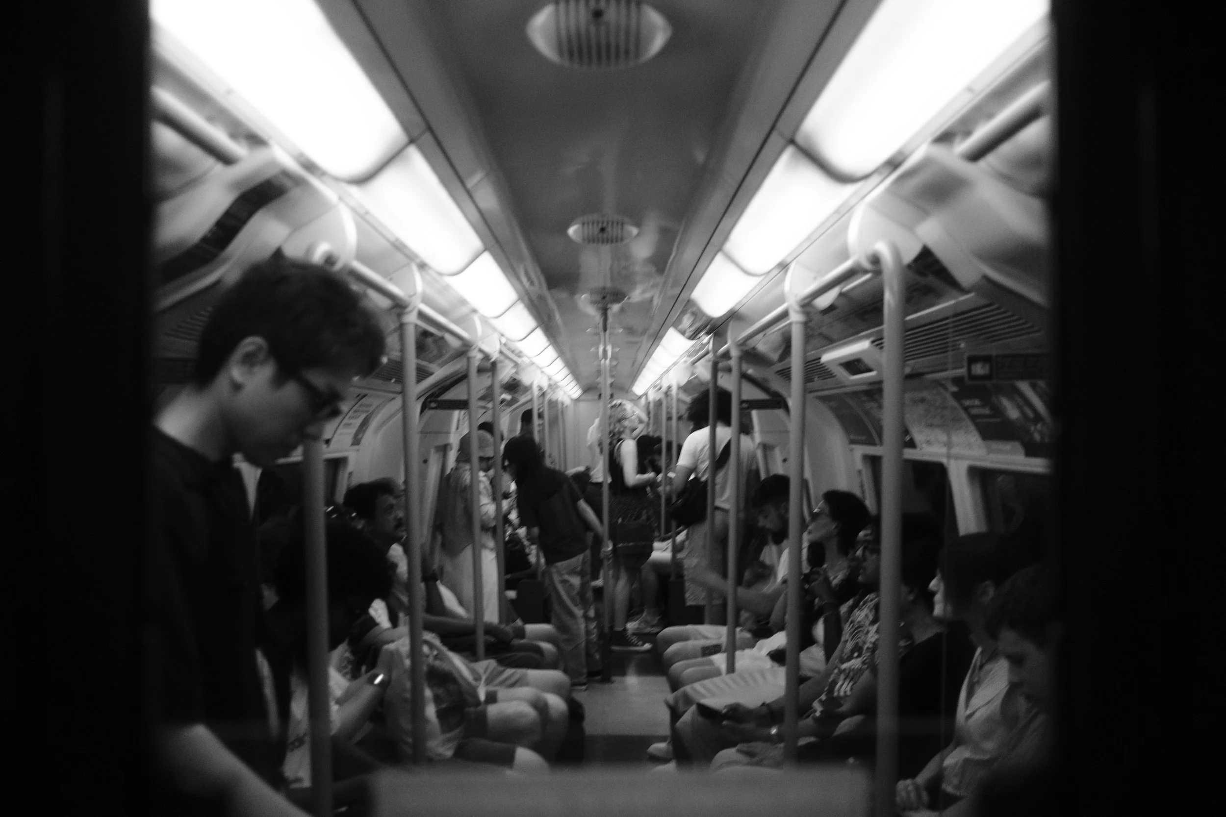Inside a subway or train car with passengers seated and standing. The image is in black and white, showing a perspective view along the aisle with overhead lights and handrails.
