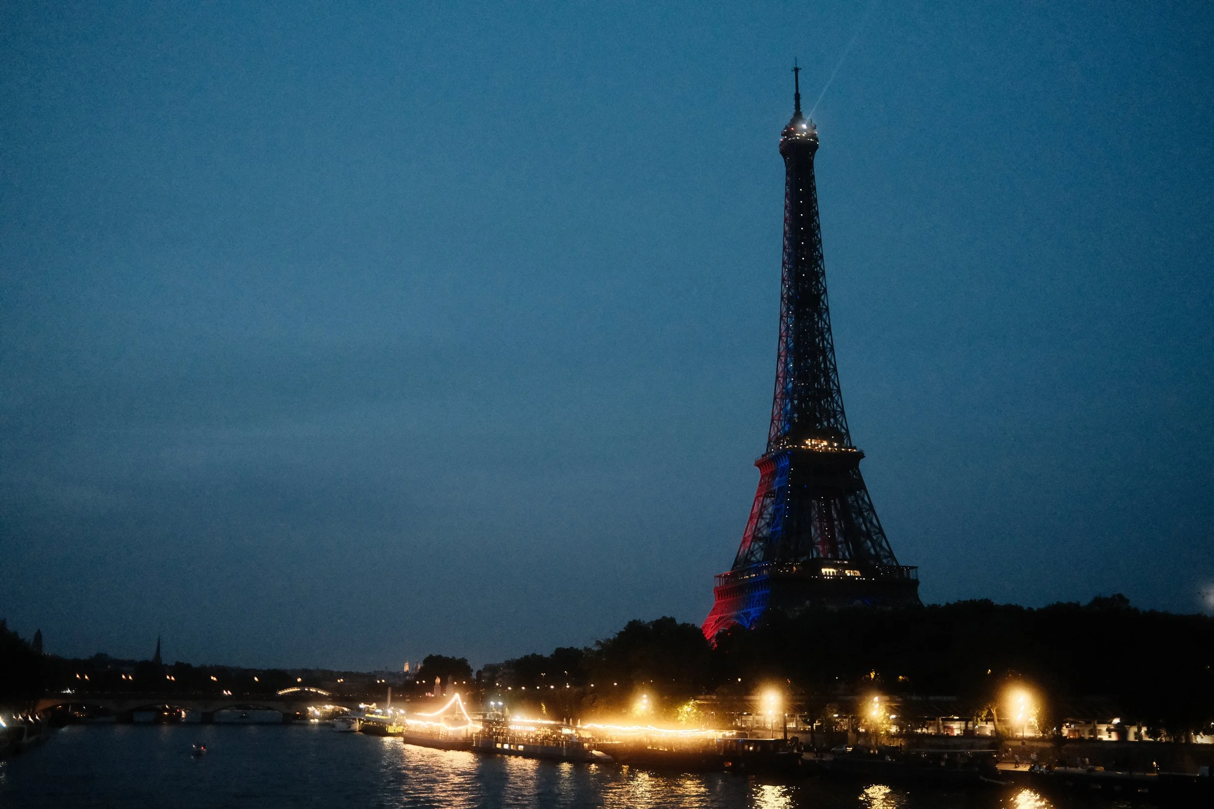 Paris, France, the Eiffel Tower illuminated at night, with lights reflecting on the river below and a clear, dark sky in the background.