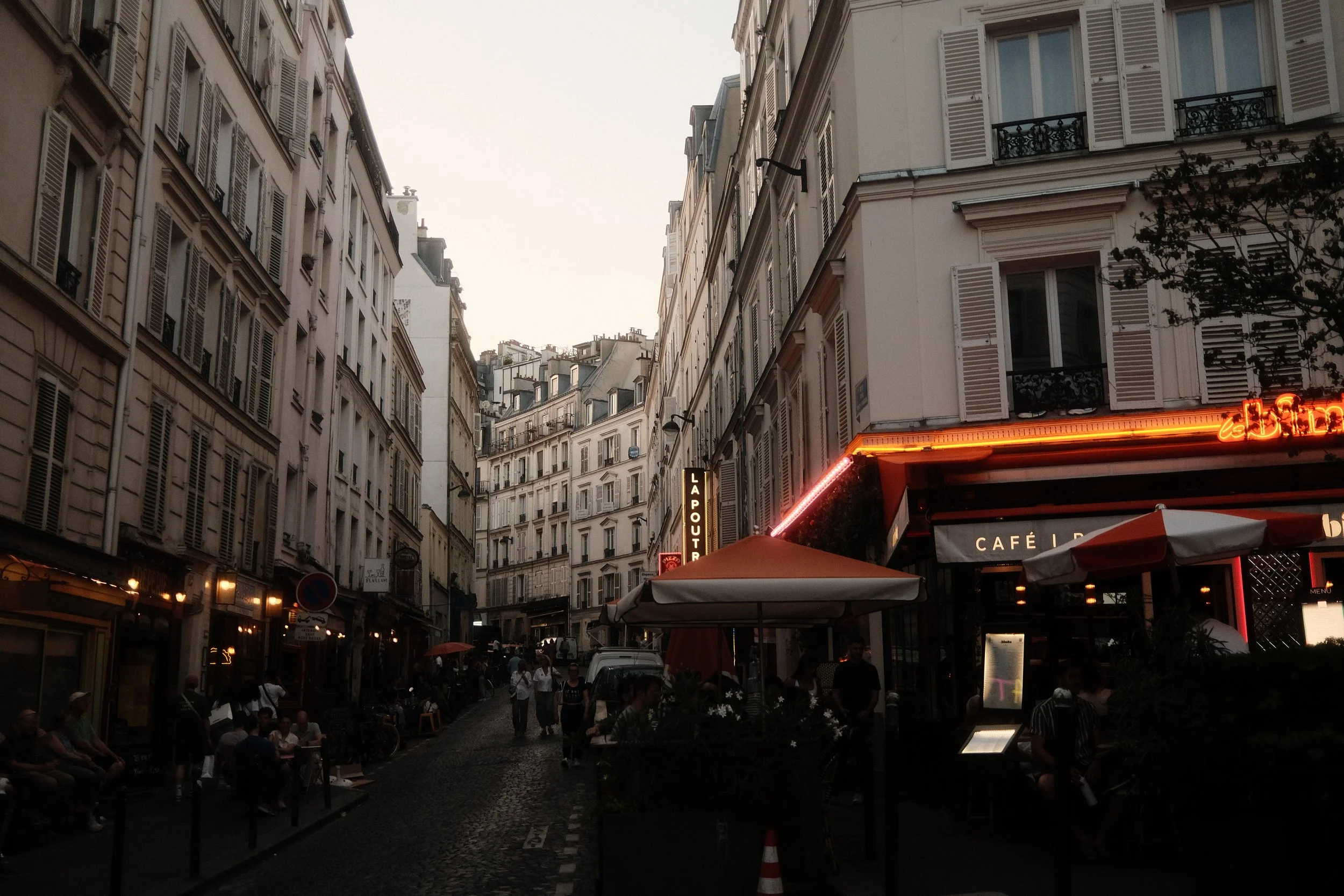 A bustling Paris street scene at dusk with tall white buildings, outdoor dining, and a neon sign for a cafe.