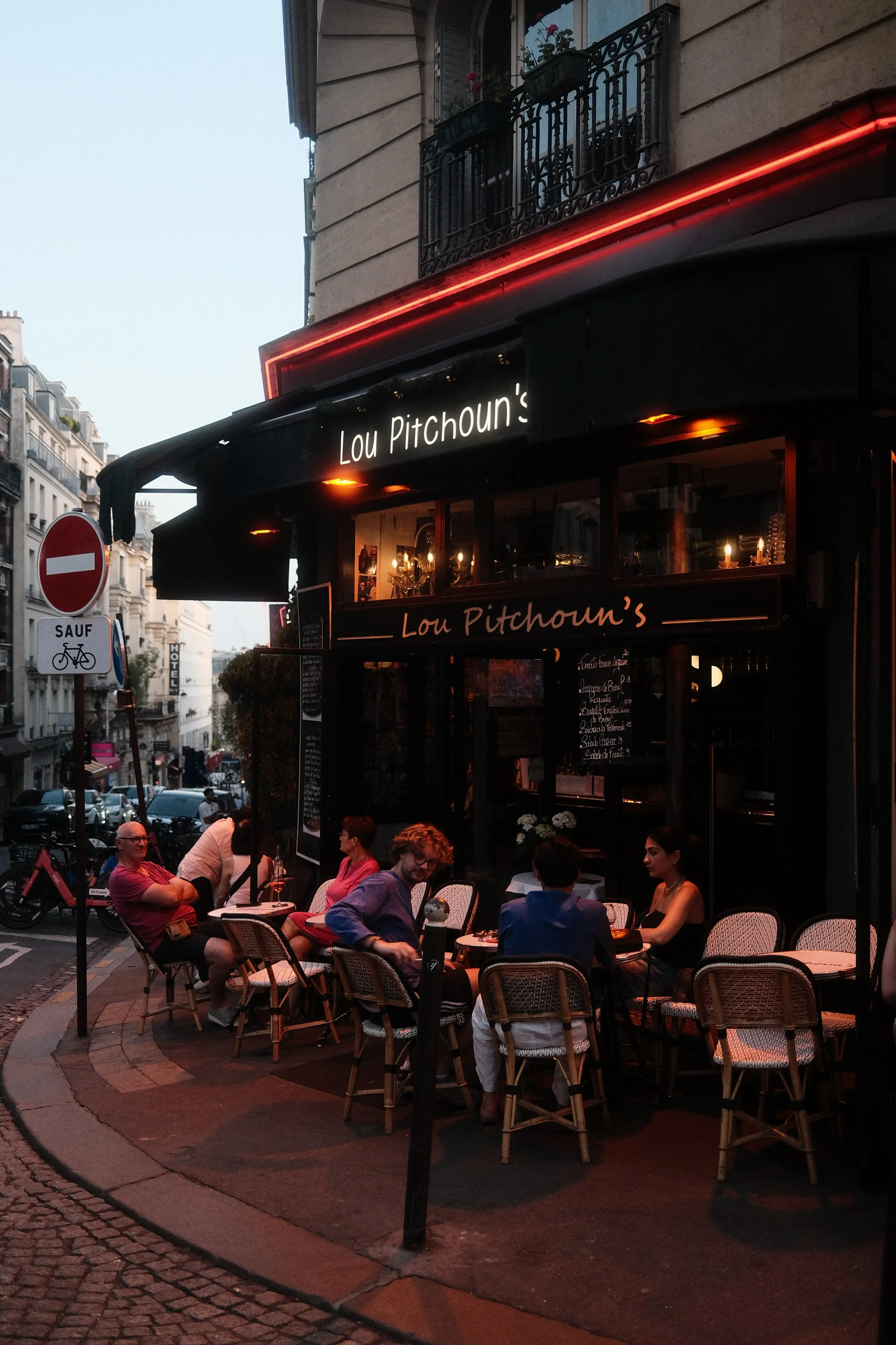 Outdoor seating area of a restaurant called Lou Pitchoun's on a city street during evening. People are sitting at small tables, dining and talking, with a building with balconies above and cars visible on the street.