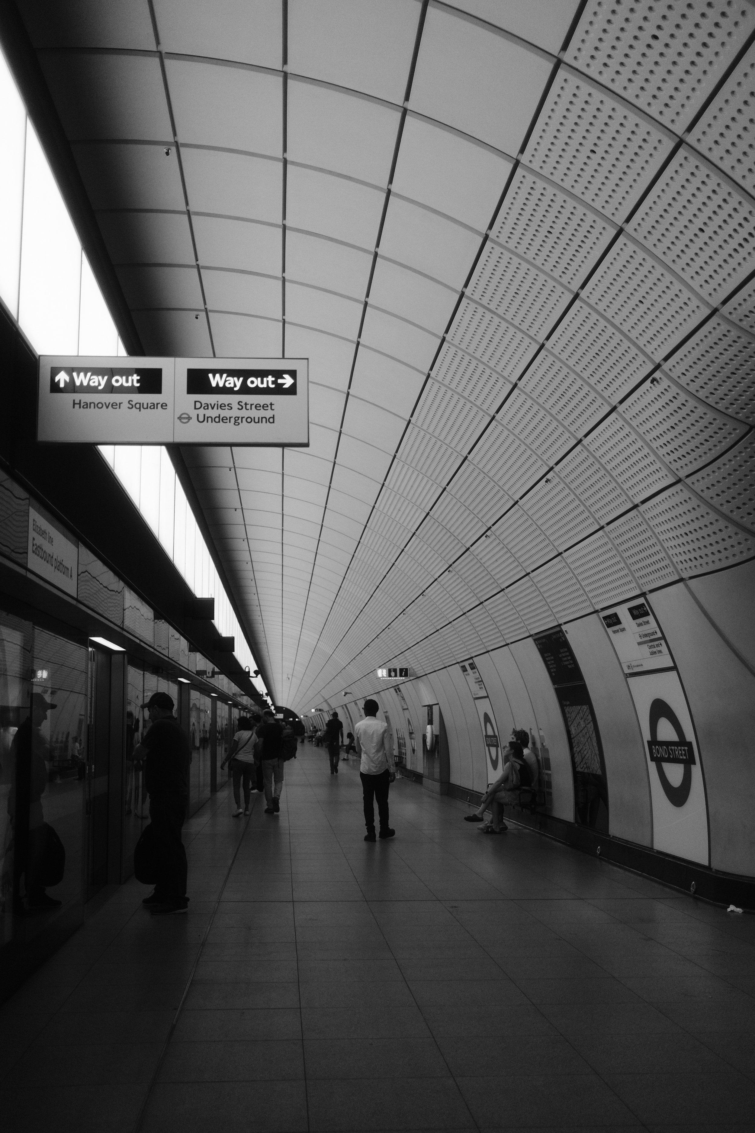 An underground subway station with people waiting and walking. Signage indicates exits to Hanover Square and Davies Street Underground. The ceiling is curved with a patterned design.