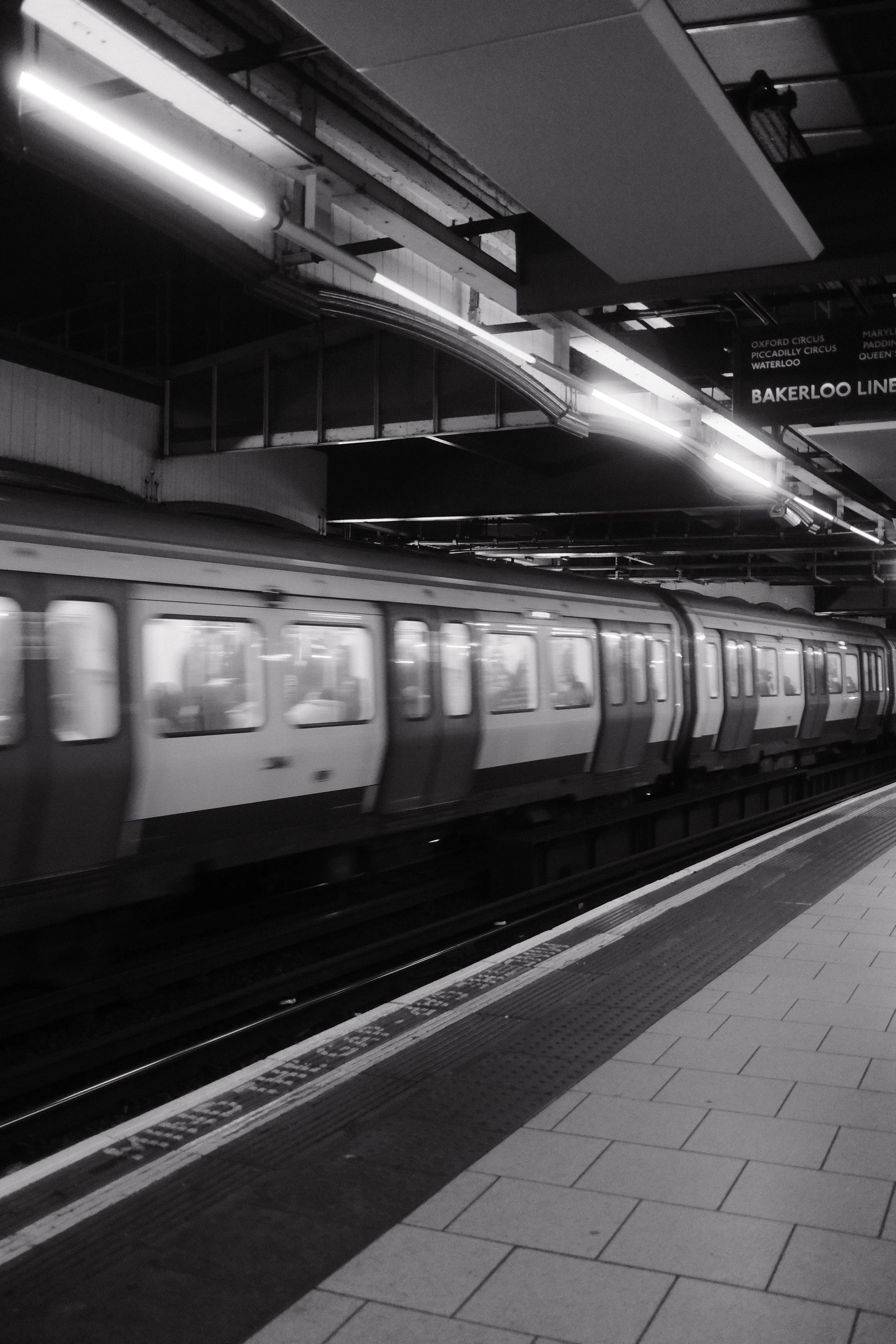 A subway train arriving at a station platform with a sign indicating Bakerloo Line, illuminated by fluorescent lights in a black and white image.
