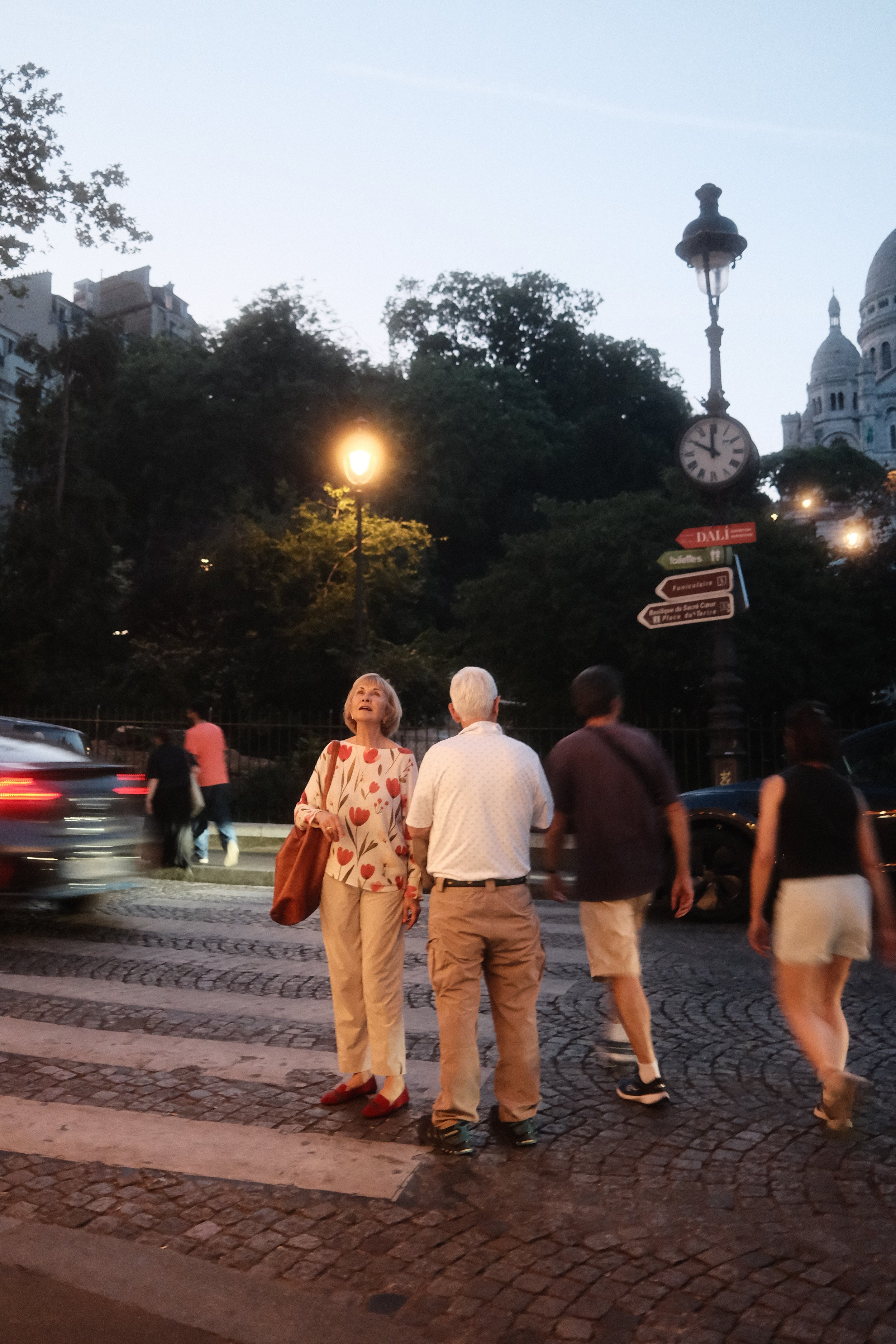 People waiting to cross a street during evening in the city, with Trocadéro building and Sacré-Cœur basilica in the background.