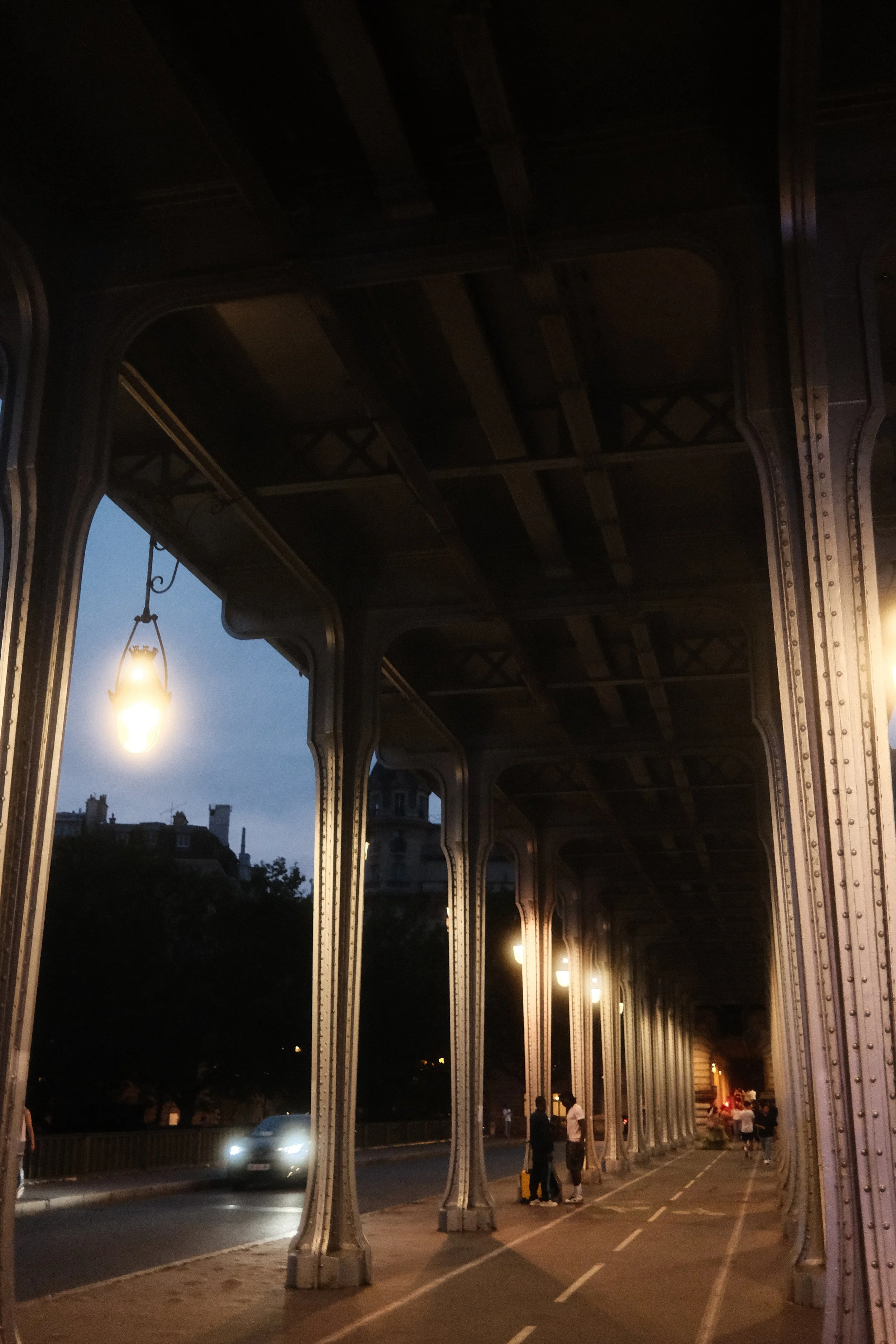 People walking and standing under a bridge at dusk with streetlights illuminating the area, city buildings visible in the background.