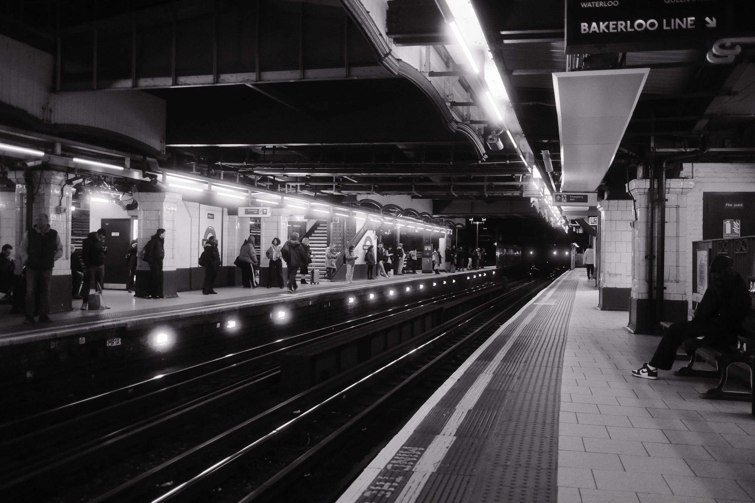 A black and white photograph of an underground subway station platform with people waiting for the train. There are signs indicating the Waterloo and Bakerloo lines, and an emergency exit sign visible. The platform has tiled flooring, benches, and li