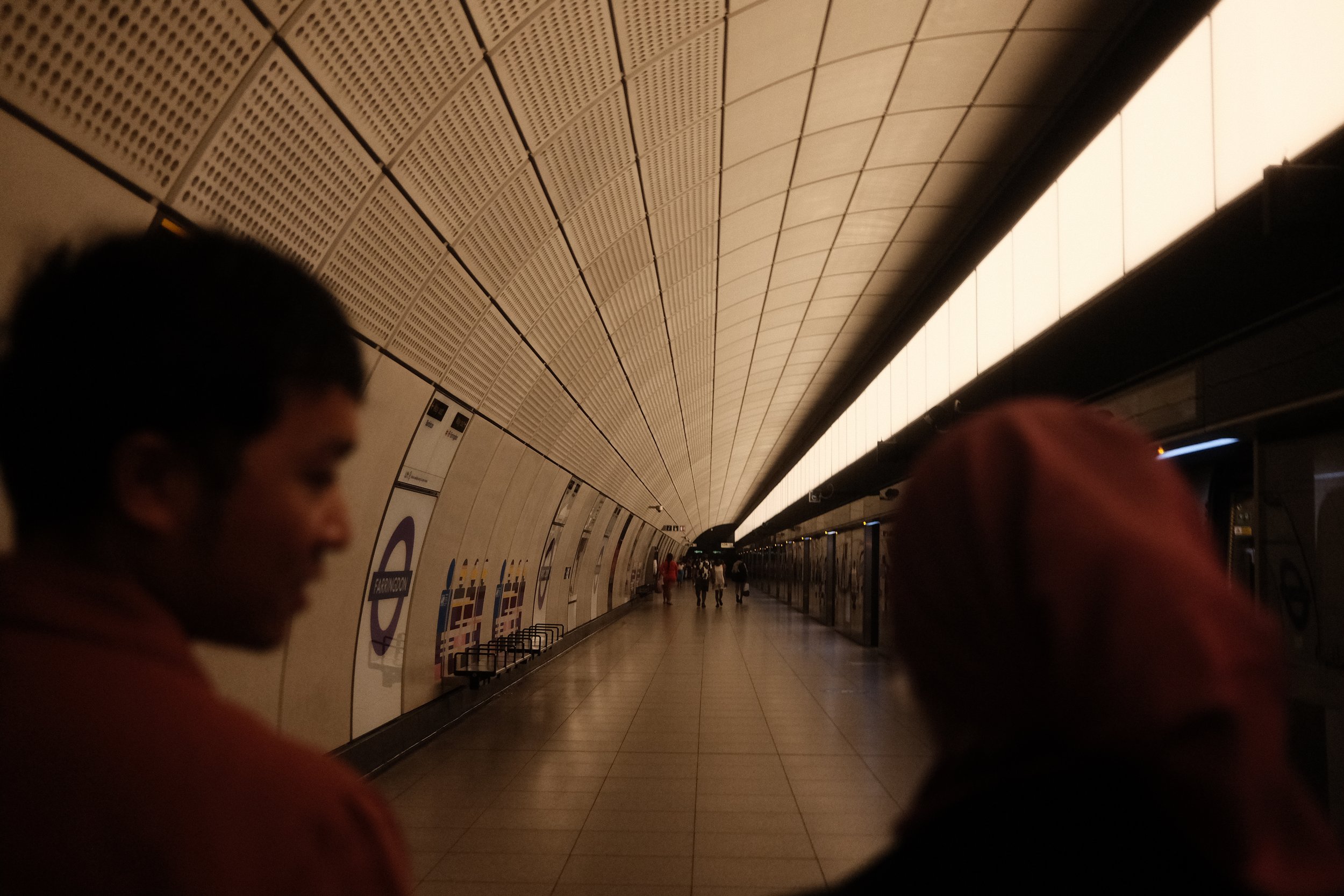 Underground metro station with curved ceiling, tiled walls, and a line of benches along the platform. People are waiting or walking in the distance.